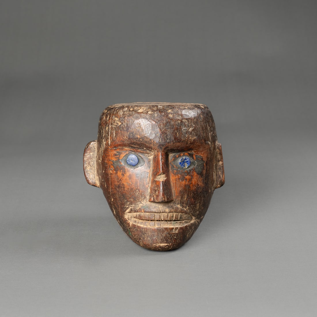 A wood Naga head: Early 20th century, the wooden head, with flattened top and back of the skull, protruding brow, nose, and ears. The eyes inlaid with blue stone, teeth inset from lips, shiny dark patina, minor loss to