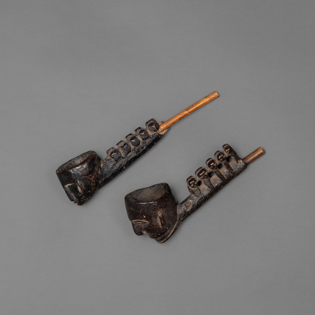 Two Naga wood pipes: Early 20th century, each pipe carved from wood, with a bamboo stem, each with a row of five heads on each side, smooth, dark patina. Lg. 7 1/2 and 7 1/4 in. Provenance: Private collection, Albuquerque