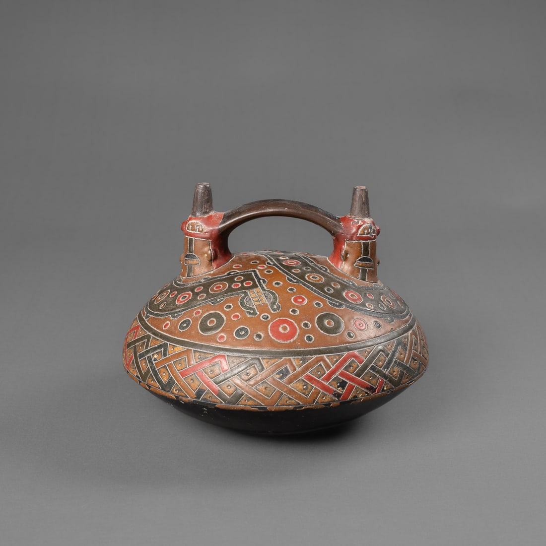 A Pre-Columbian Paracas stirrup spout vessel: Southern Peru, circa 600.100 B.C., the ceramic stirrup spout with dual cone openings, red, black, and brown post fired pigments, depicting a double-headed serpent at top. The bottom half in black pigm