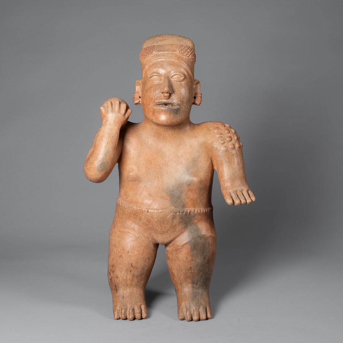 A Pre-Columbian Jalisco ball player figure: Ezatlan, circa 200 B.C. - 200 A.D., the large standing figure of a ball player with the right hand holding the ball at the shoulder, with open mount, pierced ears and wearing head gear, overall tan