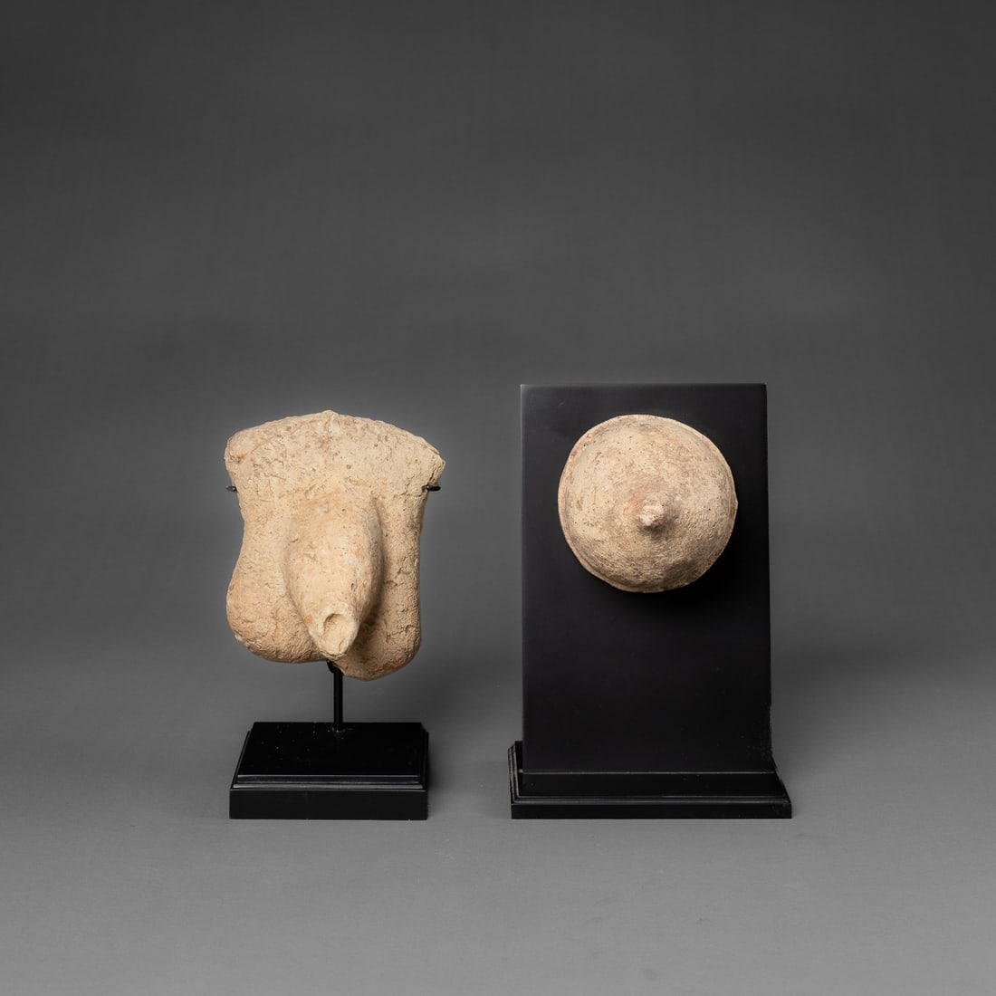 Two Etruscan votives: Circa 5th century B.C., Italy, the ceramic votives depict male and female sexual anatomy, the breast with protruding nipple, and the male pubic region with hair texture. Mounted on custom stand.