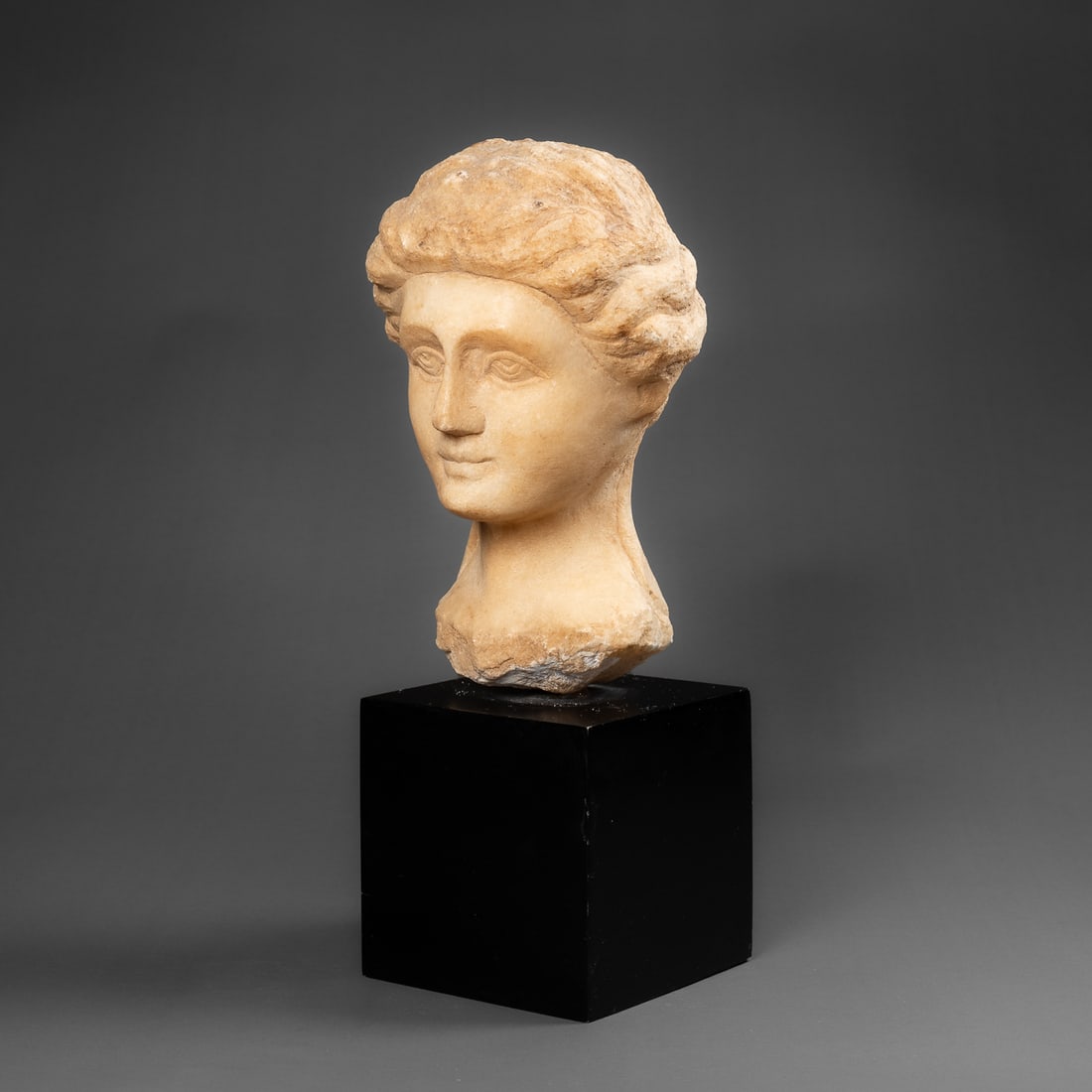 An Etruscan portrait head: Circa 5th century B.C., the stone head with detailed hair plating, tied up with ribbon at nape, the face modeled with protruding brow, careful attention to brow, eyes, and lips, ears almost covered