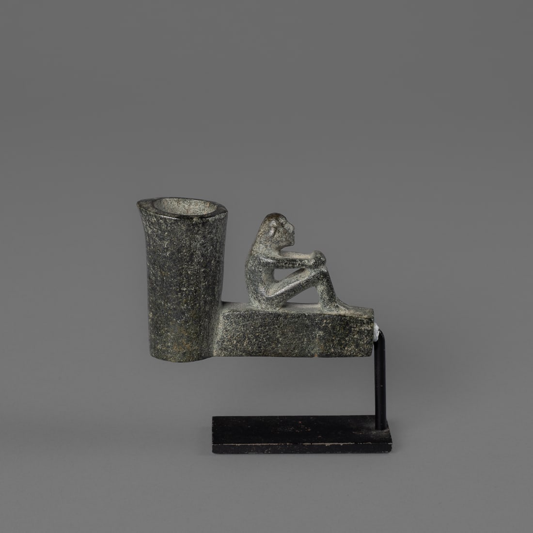Cherokee steatite effigy pipe: 19th century, or earlier, the stone pipe with square base, and long cylindrical pipe bowl, the seated figure on top of the pipe stem with arms crossed on knees, face with slit eyes, protruding nose,