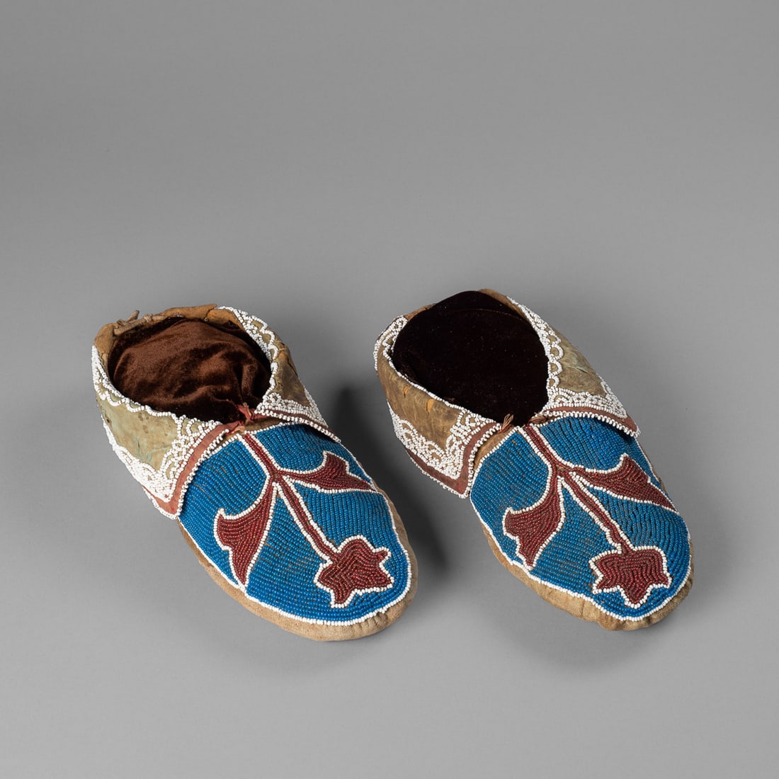 A pair of beaded Delaware moccasins: Circa 1860s, decorated with red and white abstract floral designs on a blue field, white bead work and trade cloth on the flaps, on a soft native tanned hide. Lg. 9 1/2 in. Provenance: The James