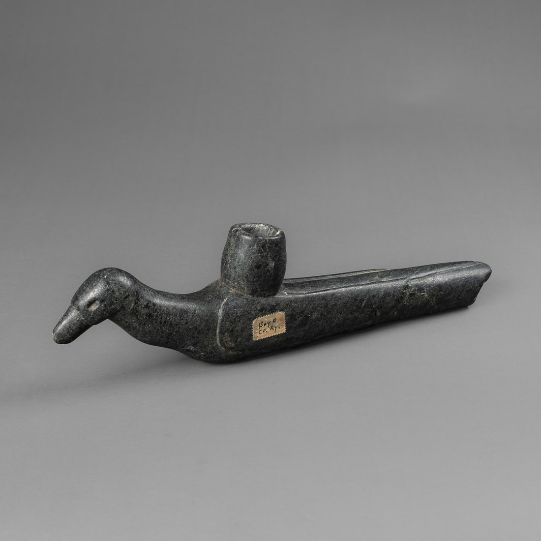 A Moundbuilder bird effigy pipe: The long black stone pipe with bird head and beak at end, carved wings with feathering on either side, tucked feet at bottom, the tail feathers at the other end with opening, pipe bowl behind head.