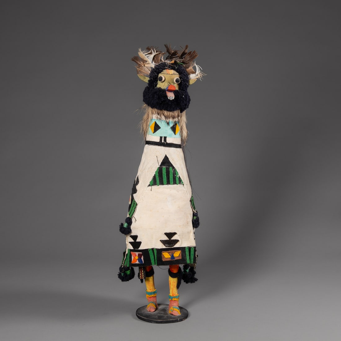 A large Zuni kachina: The tall doll with torso fully wrapped in decorated cloth garments, with black wool ruff around neck and head, long, open mouth, standing on a round wood base. Ht. 21 1/2 in.