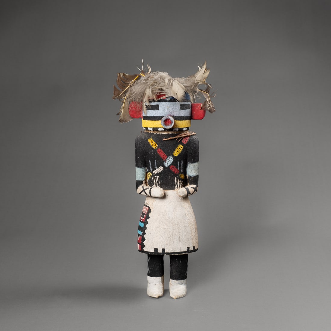 A Hopi kachina: A case mask with red ears and snout, wide black eyes, the rear with two corn devices, wearing a black shirt with colored corn across the chest, with white kilt, black leggings and white moccasins.