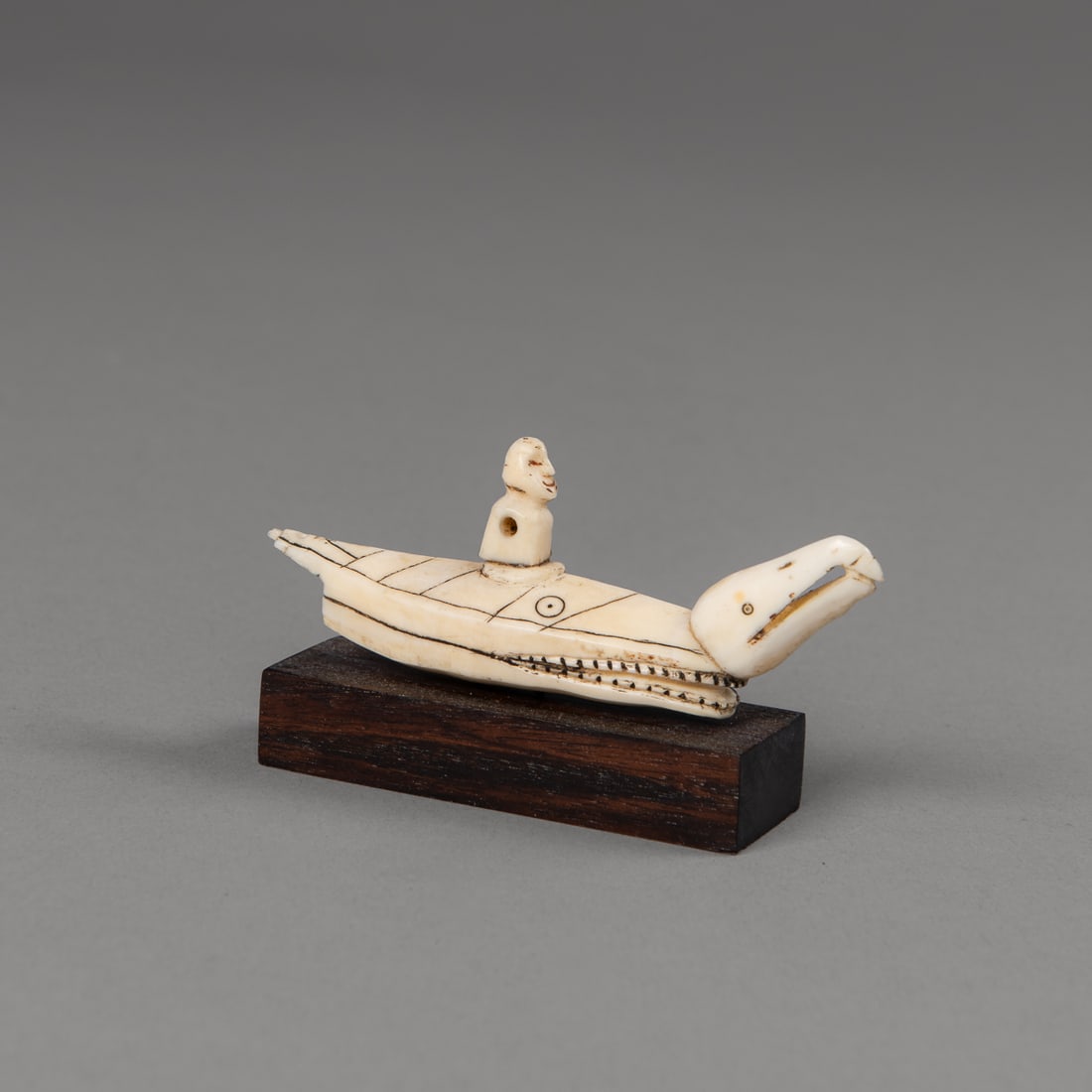 A Bering Sea Eskimo (Yupik) zoomorphic kayak Figure hunting charm: A carved wood hunting charm or amulet depicting a hunter seated in a zoomorphic, whale-form kayak, with a small bird carved at the kayak's prow. The hunter figure is carved with a conical cap; the arm