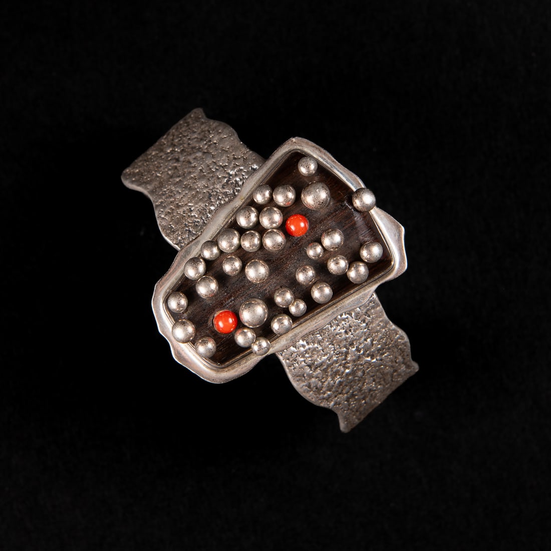 A Lyndon Tsosie cuff bracelet: Lyndon B. Tsosie (1968-), the silver cuff bracelet with textured, wavy sides, with central ebony wood setting, silver and red balls are attached by pin backing into the setting. Marked “Lyndon B. Ts