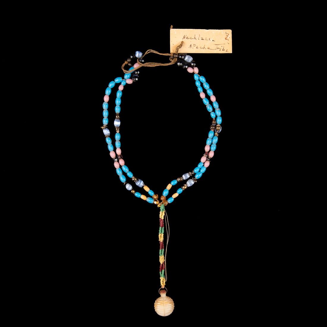 An Apache beaded necklace: Two strands to three strand necklace with red, green, yellow, blue, purple, pink, and black beads, central carved white pendant at bottom, period tag reads “Necklace Apache Tribe 1898.” Lg.