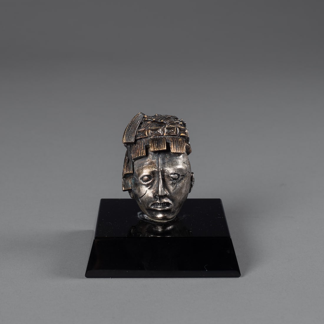 A Tane silver head: The finely modeled head, with protruding nose and headdress with layers of textured fringe, on custom stand that reads, “Head from Palenque, Mayan Culture Classic Period, 500-800 AD. Stucco head