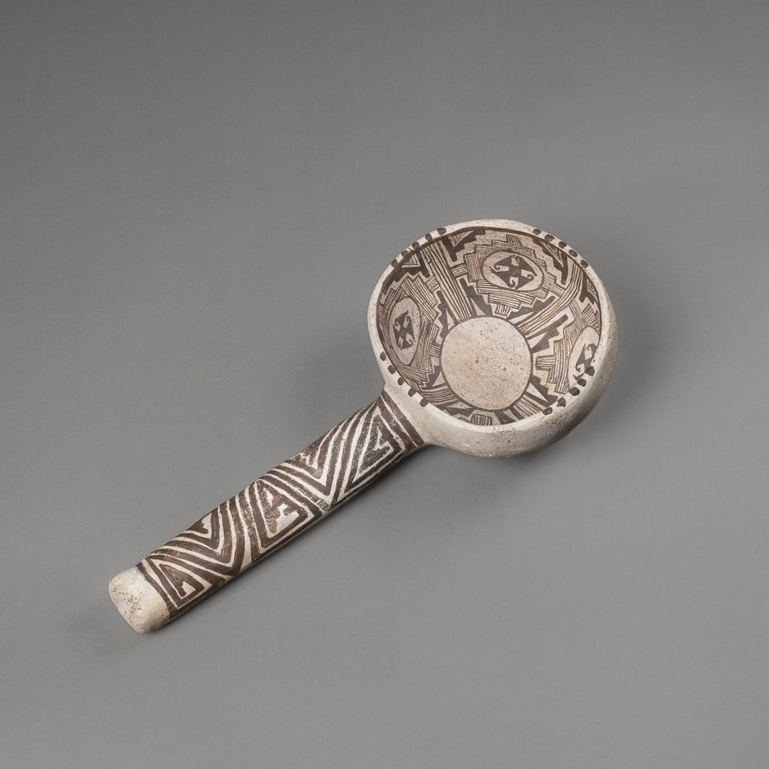 An Anasazi black-on-white dipper: Puebloan, circa 1100 AD, the black-on-white ceramic with geometric triangular patterning on handle with two feet-like protrusions at the end, rattle inside, inside the convex bowl a central circle