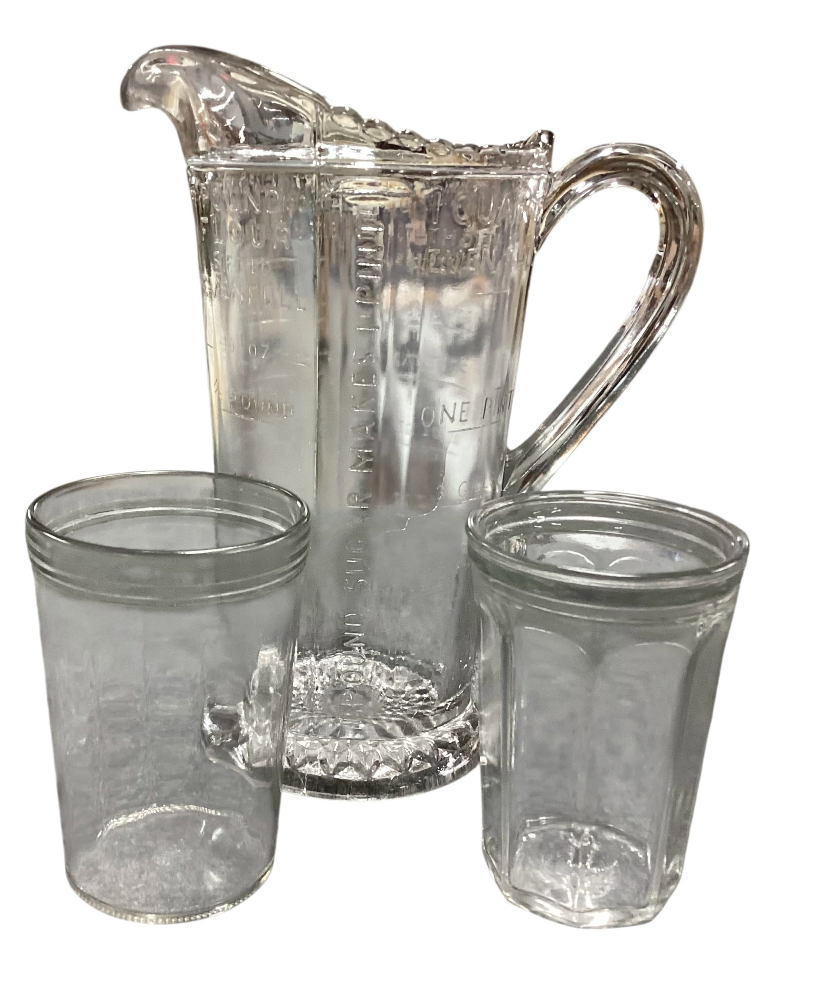 Depression-Era Glass Measuring Pitcher with Two Drinking Glasses: Depression-era clear glass measuring pitcher with handle and molded measurement markings, accompanied by two clear drinking glasses. Pitcher markings include --Good Cooking Starts with Accurate Measur