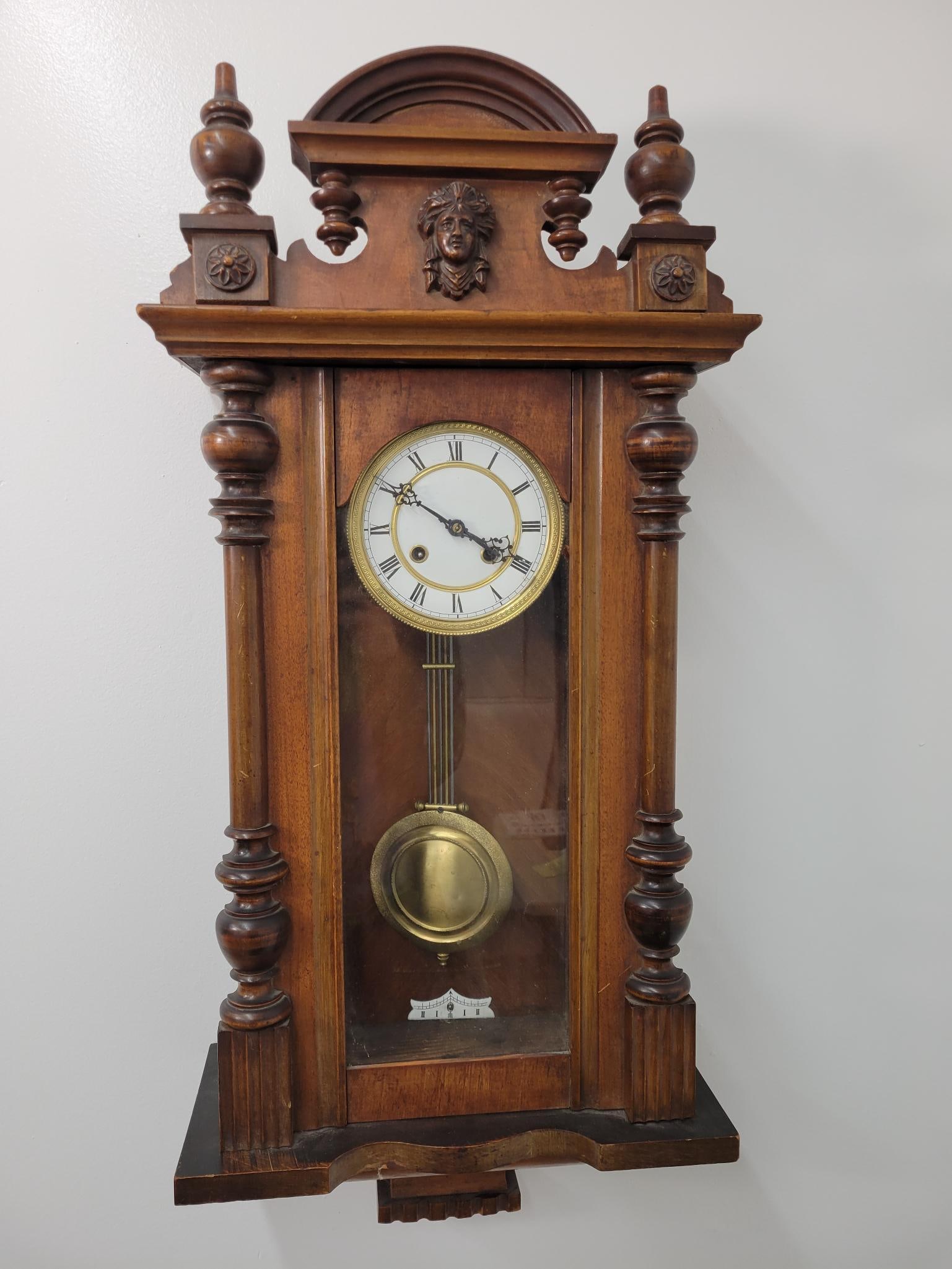19t C. Vienna Wall Clock: This lot includes a turn of the century Vienna wall clock. The clock includes the original pendulum & key and is missing some of the finials from the top and bottom of the clock. The crown piece is lo