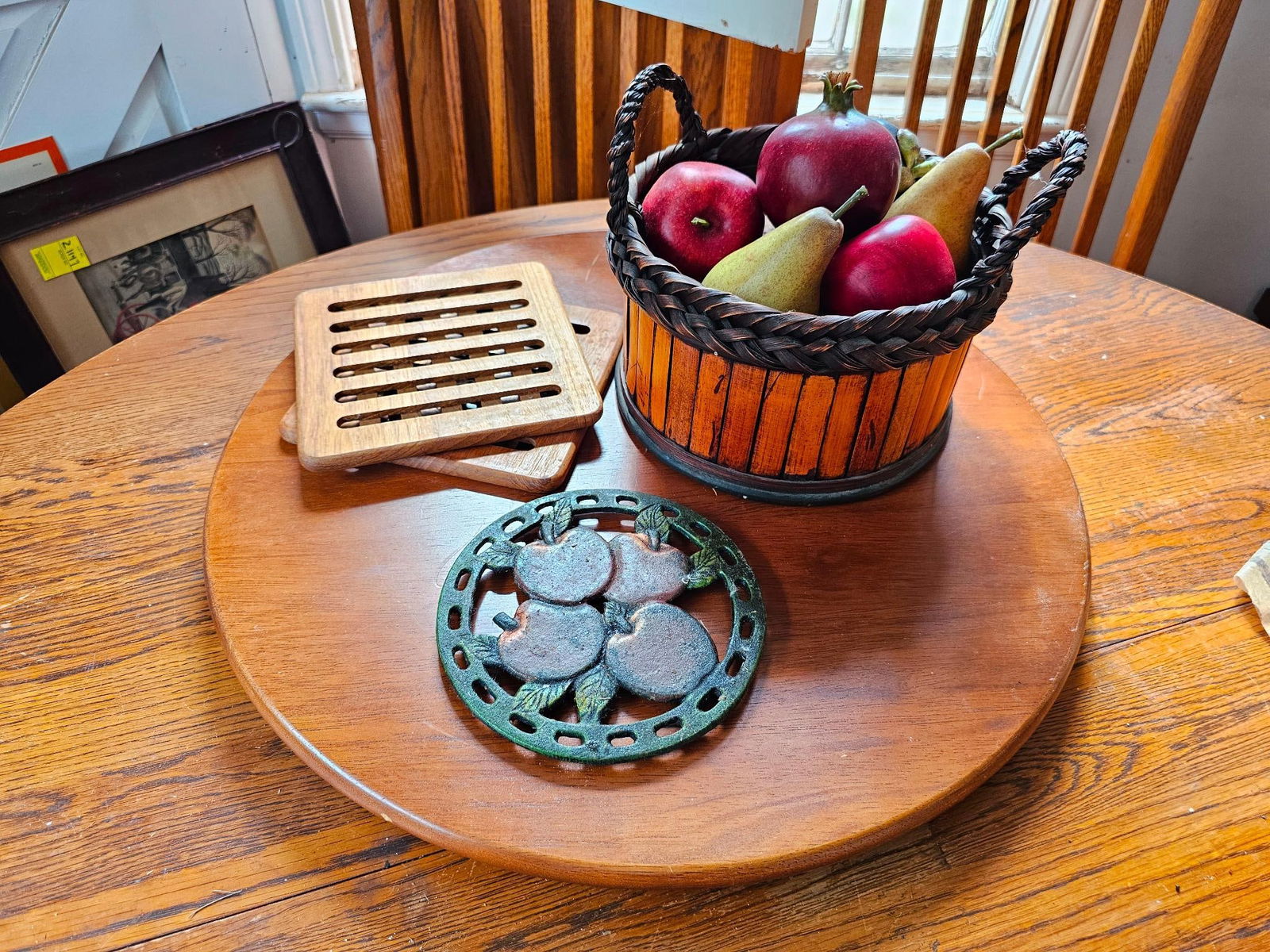 Basket with Fruit Lazy Susan Wooden & Iron Trivets: A collection featuring a woven basket with fruit, two wooden trivets, and a cast iron trivet with an apple motif all atop a wooden lazy Susan. This lot is offered in estate-found condition. Conditions