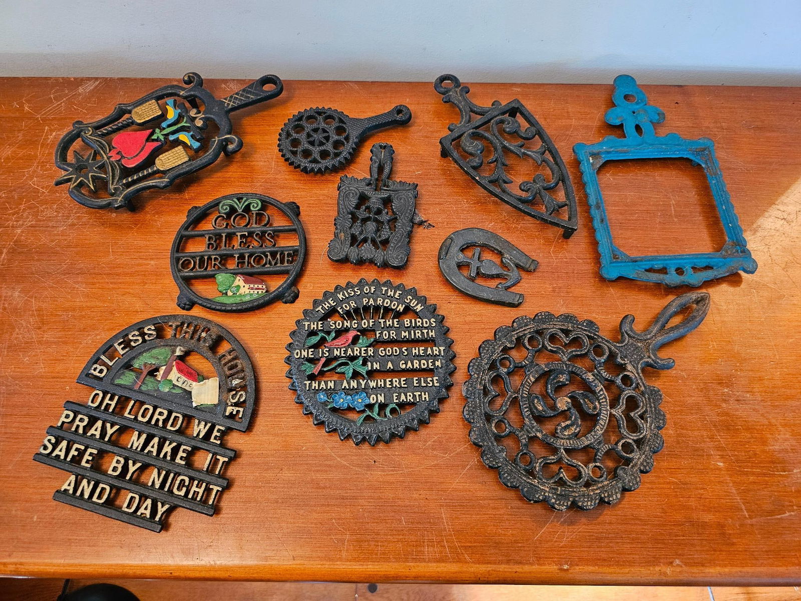 Collection of Cast Iron Trivets: Assorted cast iron trivets, featuring religious blessings and decorative designs. Some show colorful painted details. Offered in estate-found condition. Please refer to photos for more details.Measure