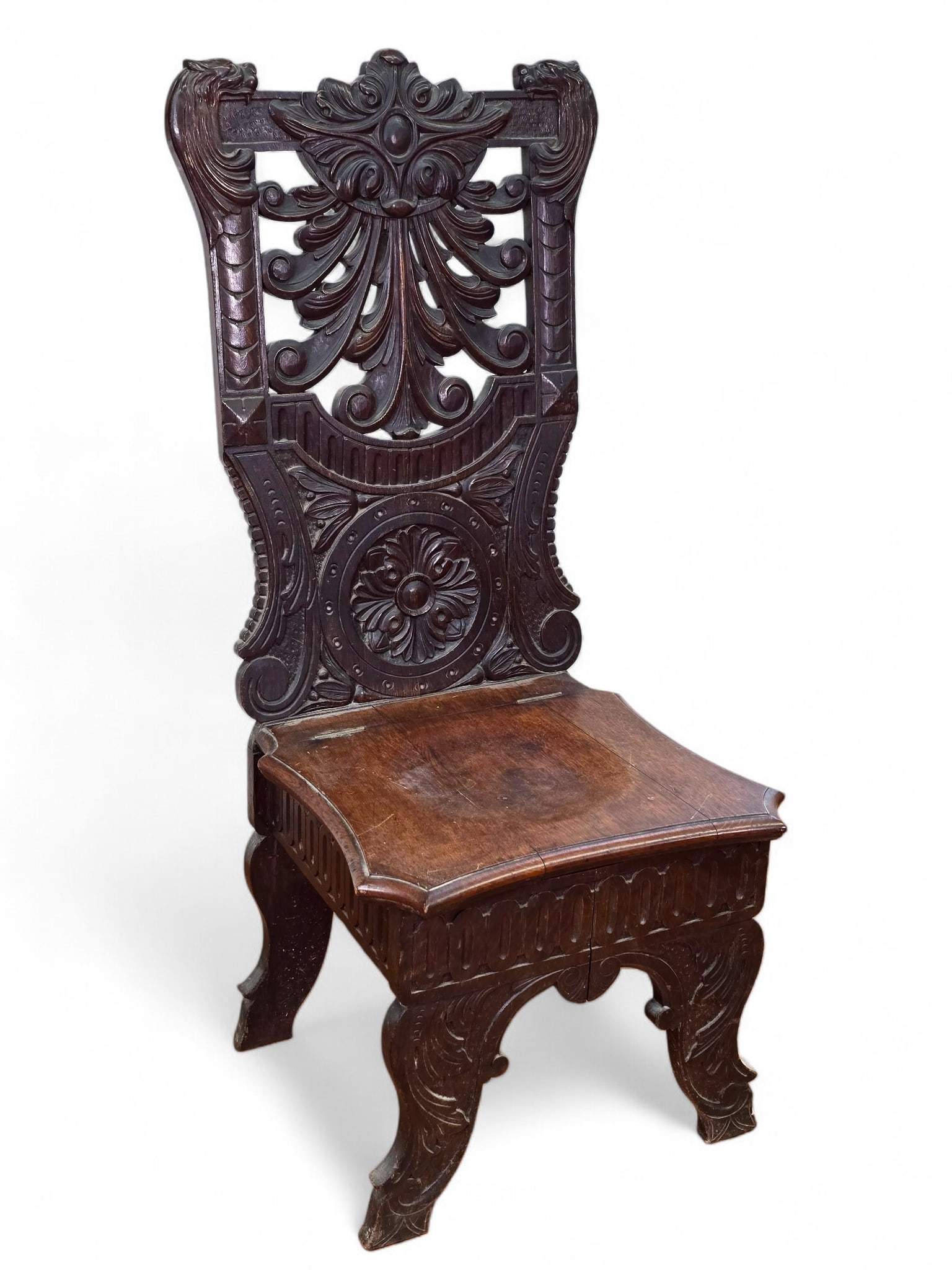 18th C. Carved Oak Commode Chair: A carved oak commode chair with an intricately carved back featuring lions and scroll designs and a paneled hinged seat that lifts. Surface wear and natural splits consistent with age and use. See pho