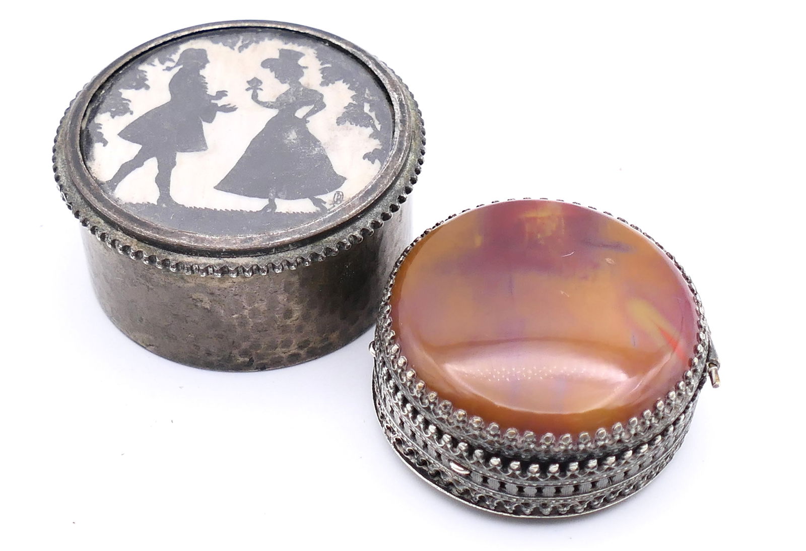 Two Antique Snuff or Pill Boxes: Two antique snuff or pill boxes, one with a repousse hinged lid depicting a park scene with a stone cabochon lid, alongside one with a courting couple with a hammered lid and silverplate body. Size: L