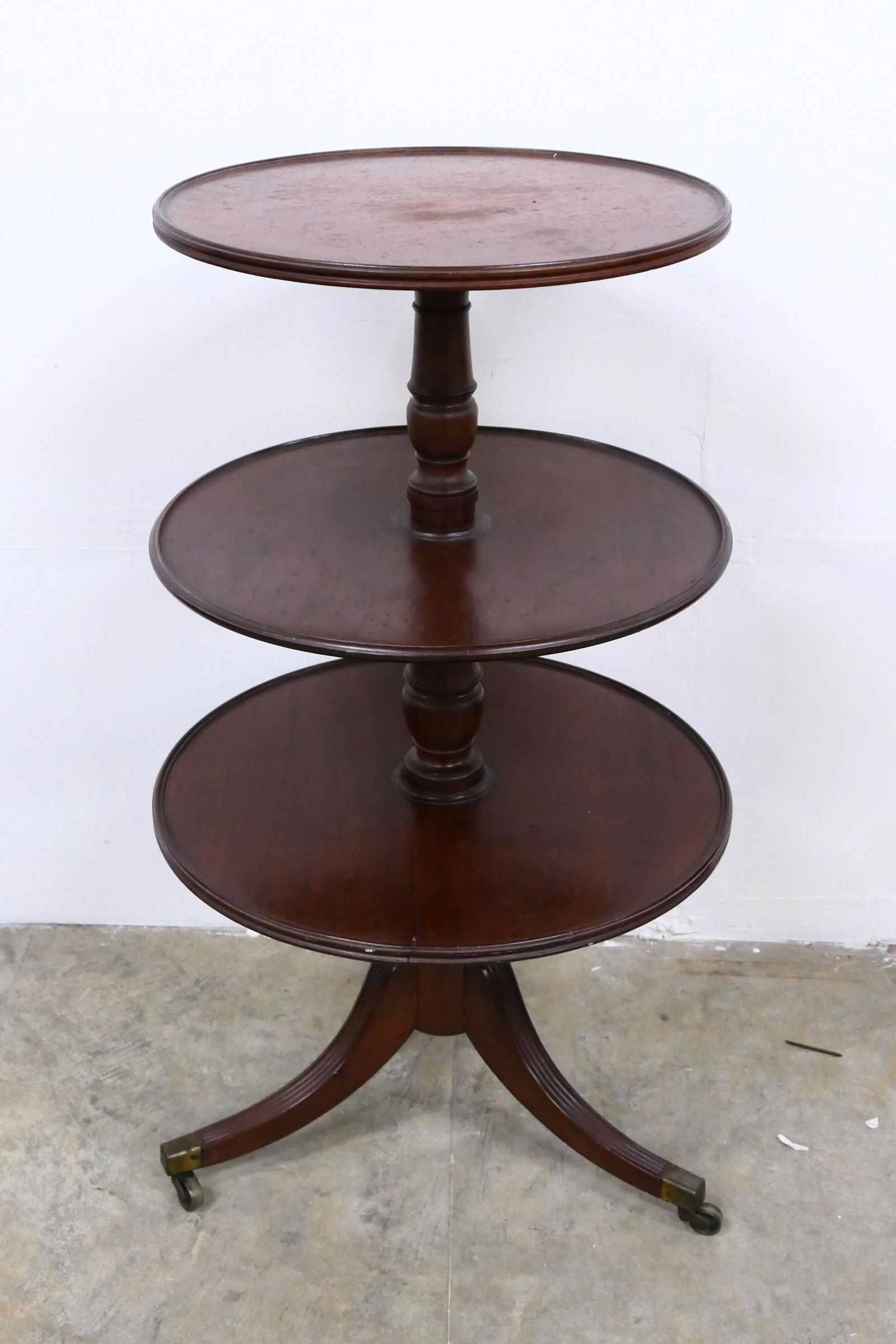 Vintage Mahogany Style Dessert Table: Circa 1940 mahogany finish Duncan Phyfe style triple-tier circular dumbwaiter or dessert table with short gallery edges, turned post, and trefoil legs with brass capped feet and casters. The three tie