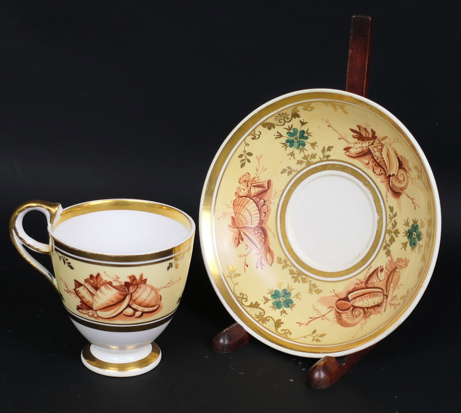 Antique English Davenport Teacup and Saucer: Antique English Davenport porcelain teacup and saucer featuring a peach field with seashell and clover border. The cup handle is adorned with a figural face peering into the cup. Teacup bottom shows a