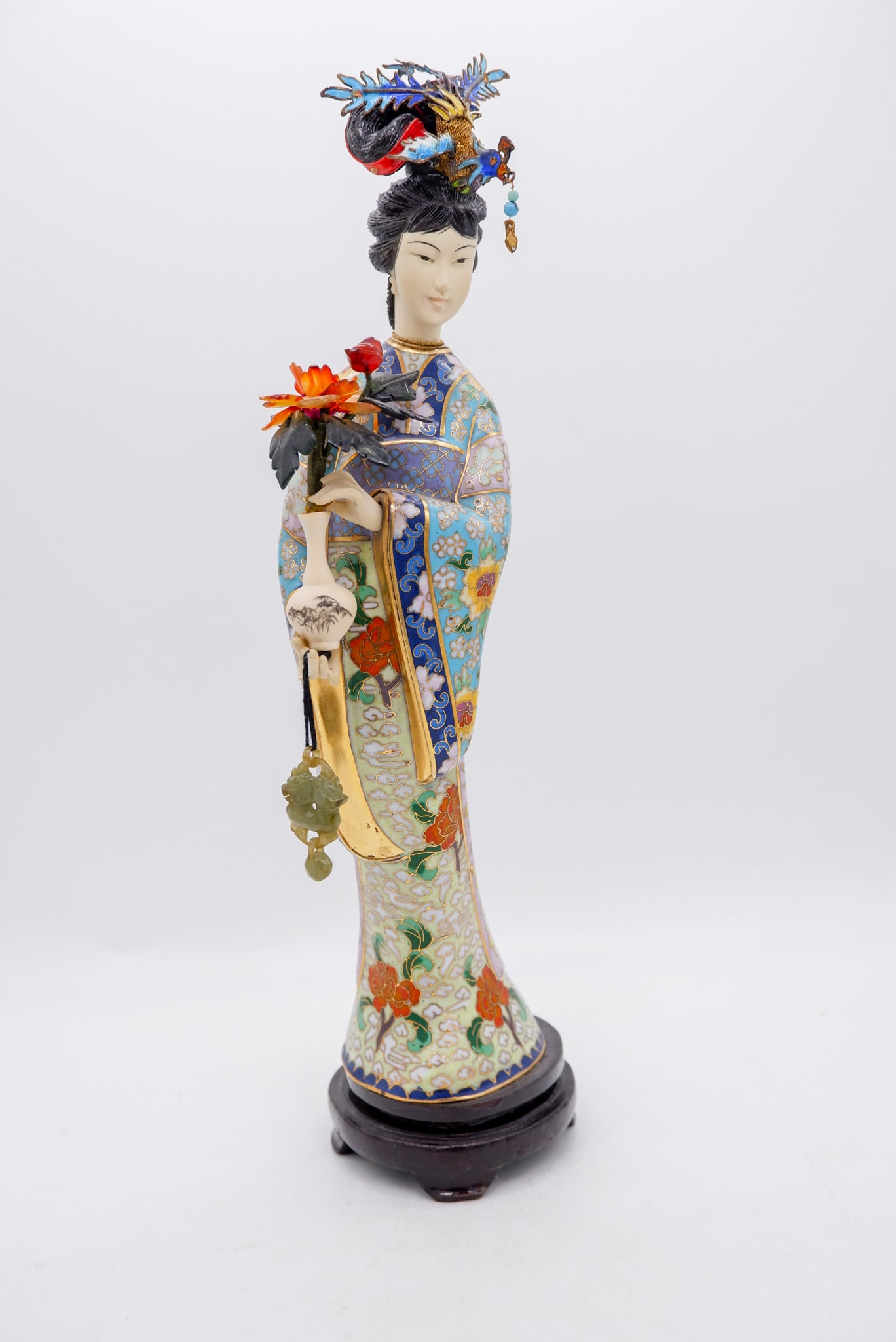 Cloisonne Figurine of a Geisha in Traditional Attire: Cloisonne figurine of a geisha in traditional attire holding a vase of flowers and adorned with elaborate headwear featuring a bird, on attached wooden base. Size: 14"h Location: Sitting Room