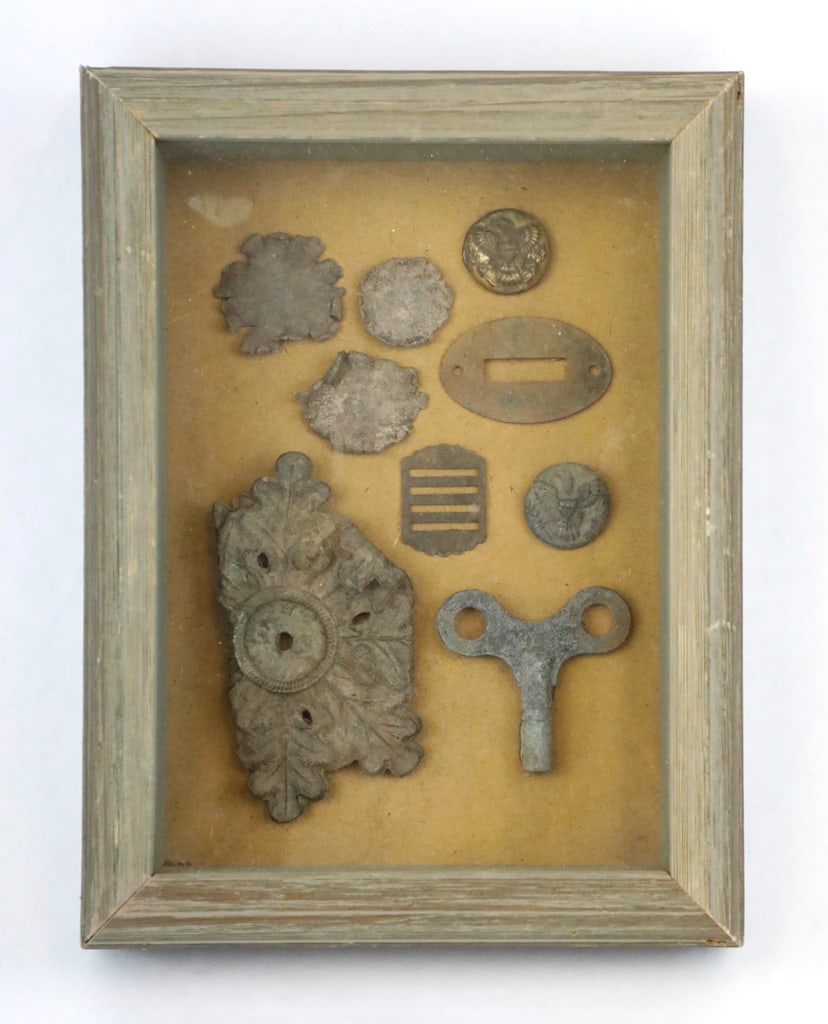 Collection of Metal Relics: Collection of metal relics, including: two round buttons with embossed designs depicting eagles, a dress helmet plate, clock key, and more. Displayed in a wood shadowbox frame. Size: 6.25"w x 8.25"d x
