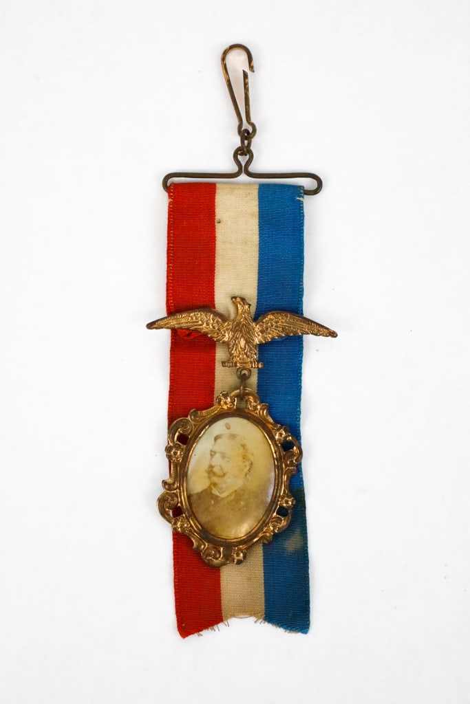 Presidential Campaign Collectible of William H. Taft with Eagle on a Ribbon and Clip: Pressed metal framed photo of William H. Taft in ornate frame suspended from a red, white, and blue ribbon on a brass-tone clip. Show discoloration from age. Size: 5.25"l
