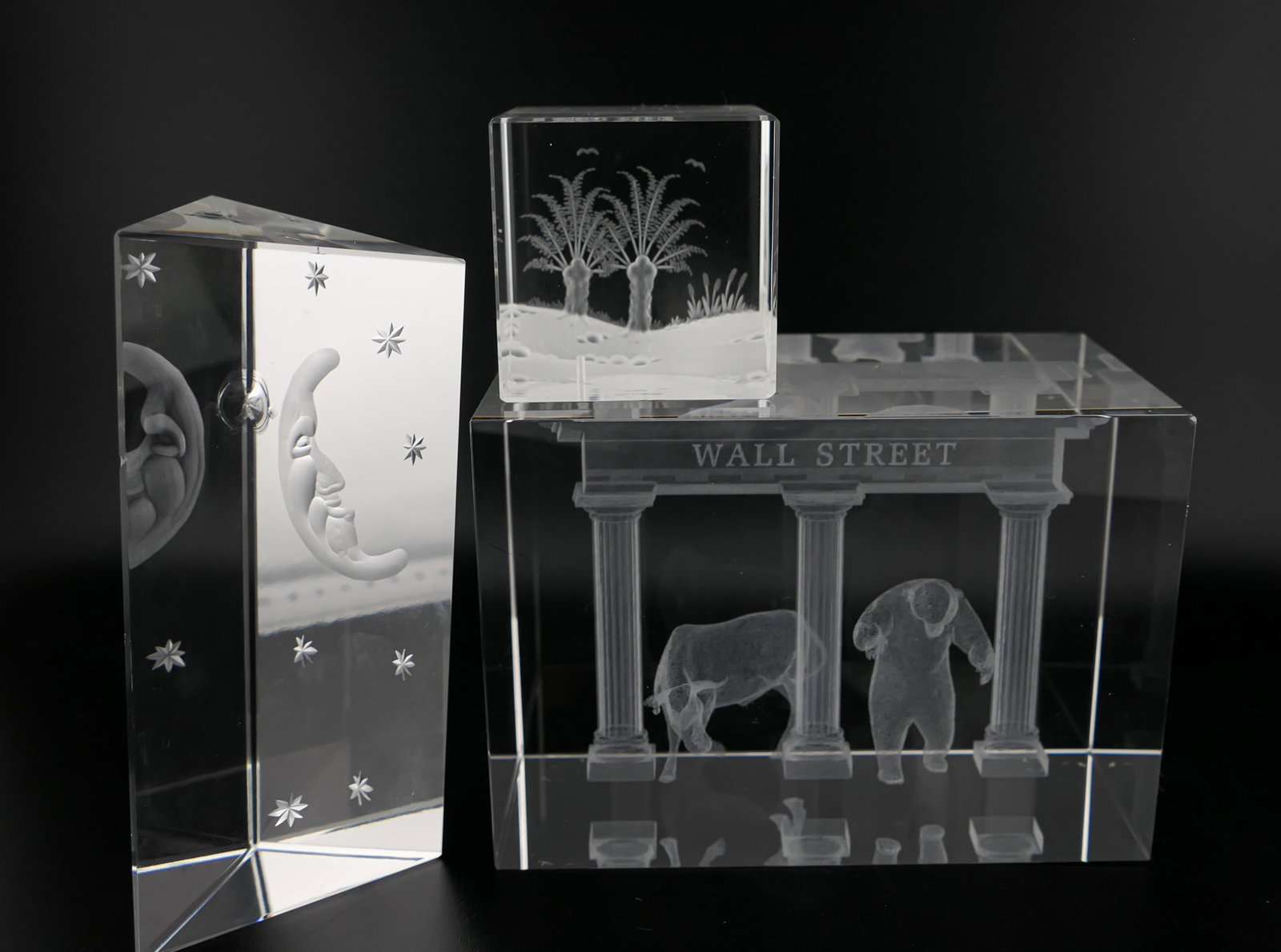 Geometric Crystal Accent Pieces: Geometric crystal accent pieces including a triangular paperweight etched with a crescent moon face motif (Marked TNECC90), a Steinbach laser-cut Pine Hill cube, and a Wall Street bull and bear laser 