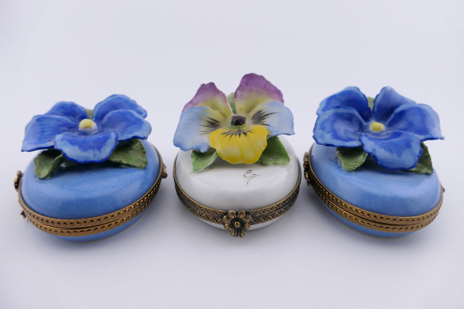 Three Limoges Porcelain Pansy Trinket Boxes: Marked Limoges hand painted porcelain hinged trinket boxes, each featuring an applied pansy motif on the lid, one of which is colorful, two of which are blue with gilt accenting. All are marked on the