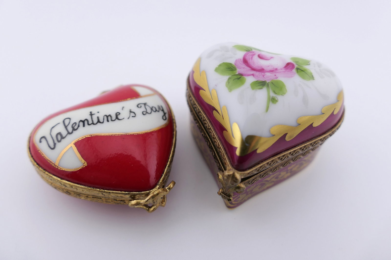 Two Limoges Porcelain Heart Trinket Boxes: Two marked Limoges hand painted porcelain hinged trinket boxes featuring heart shapes with red, burgundy, and gilt accenting. The red box contains feather and heart trinkets. Both are marked on the bo