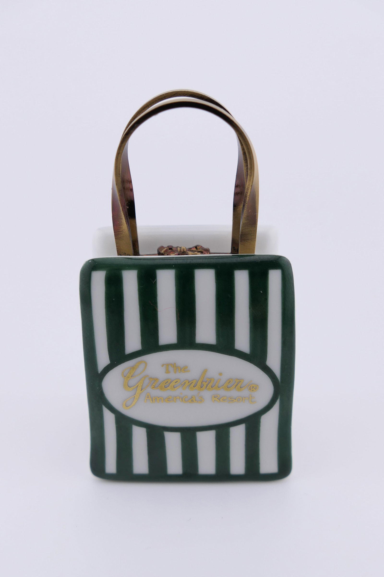 Limoges Porcelain Greenbrier Resort Trinket Box: Limoges hand painted porcelain hinged trinket box featuring a gift bag design branded for The Greenbrier Resort, and containing a small present trinket. Marked on the inside Limoges France. Excellent 