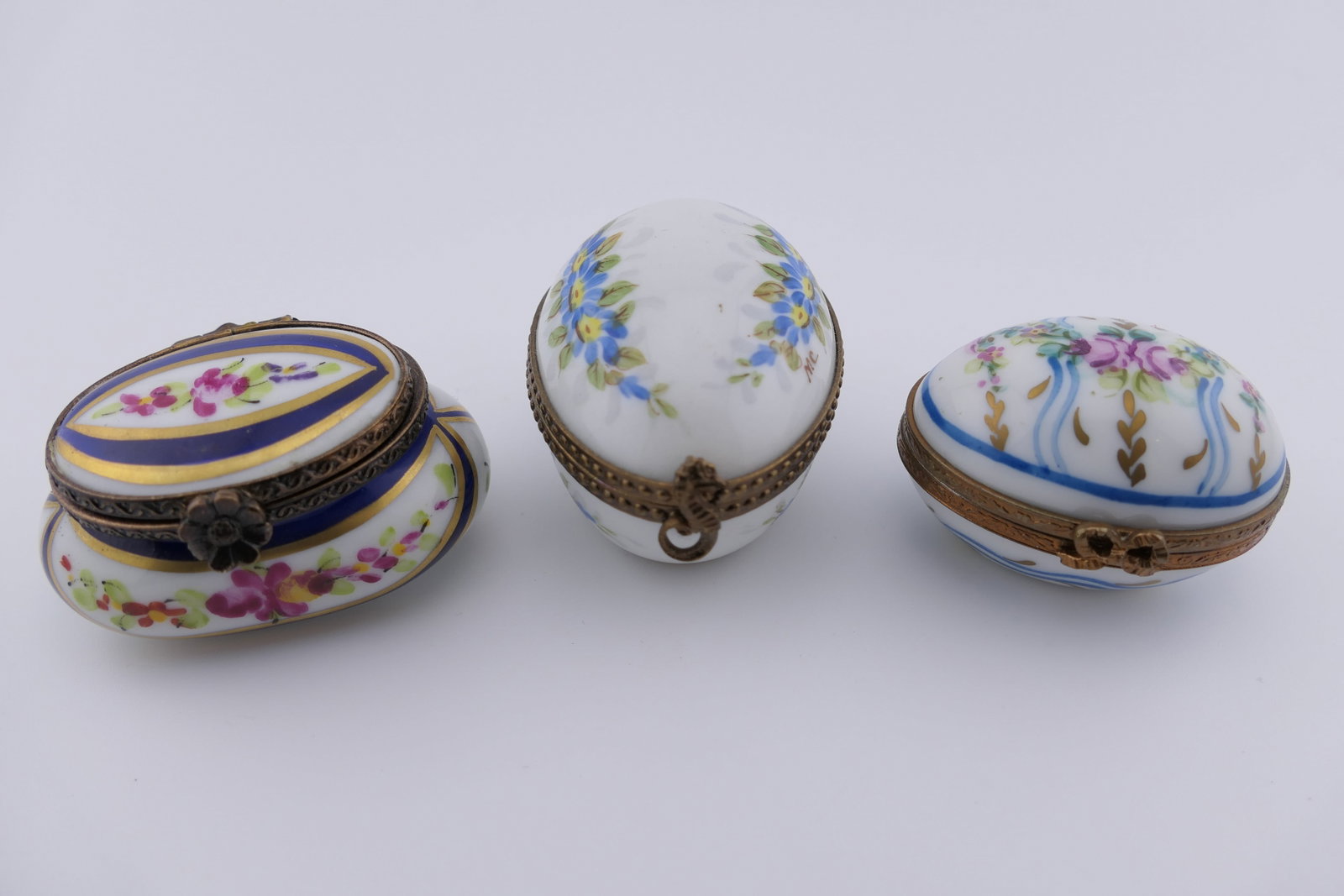 Three Limoges Porcelain Trinket Boxes: Three marked Limoges hand painted porcelain trinket boxes including an egg-shaped trinket box with floral motifs and two floral trinket boxes, one of which is egg-shaped, the other is cushion-shaped. 