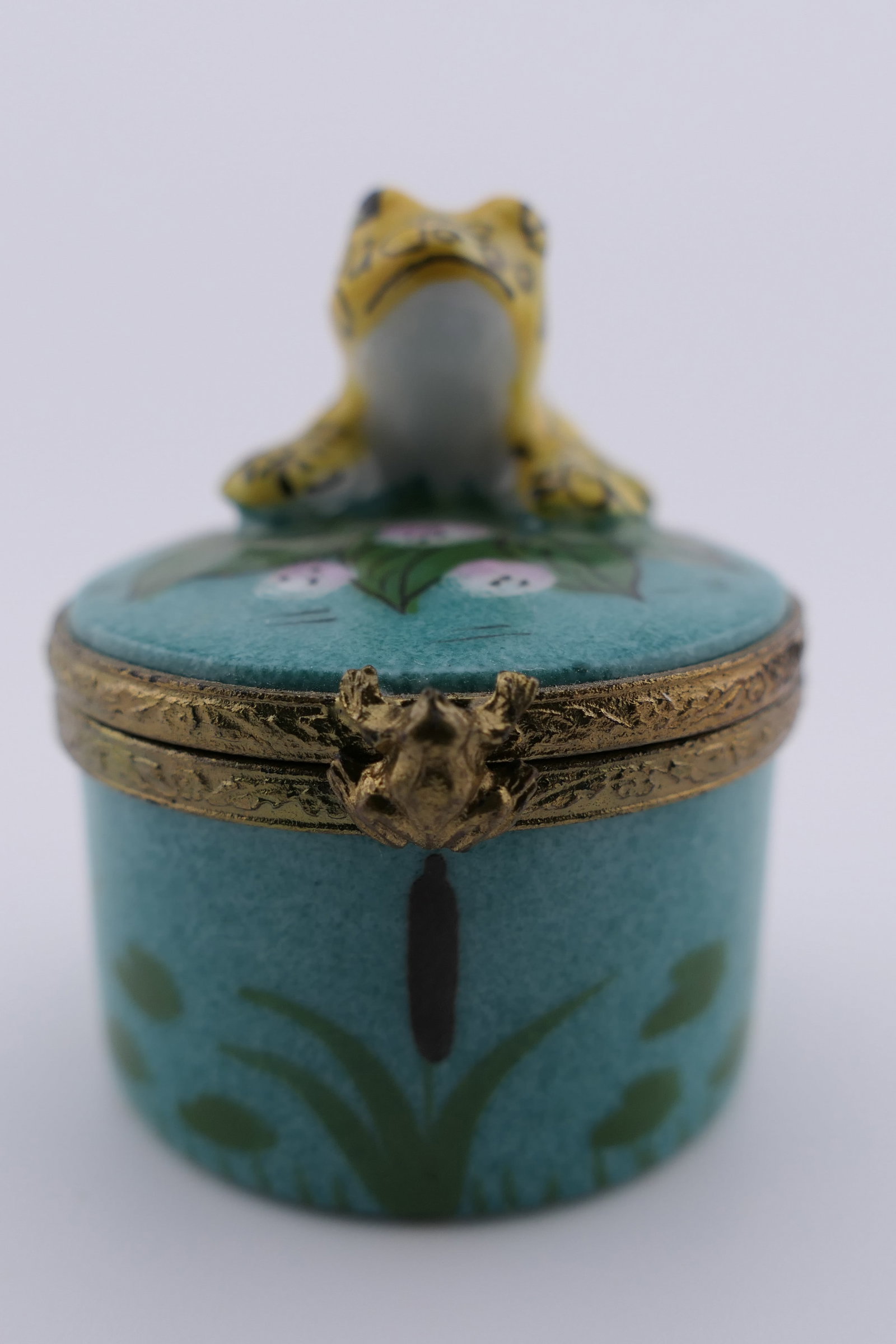 Limoges Porcelain Frog Trinket Box: Limoges hand painted porcelain hinged trinket box featuring an upper design of a yellow frog over cattail motifs. Marked on the bottom Limoges France. Excellent condition. Size: 2"h Location: Living R