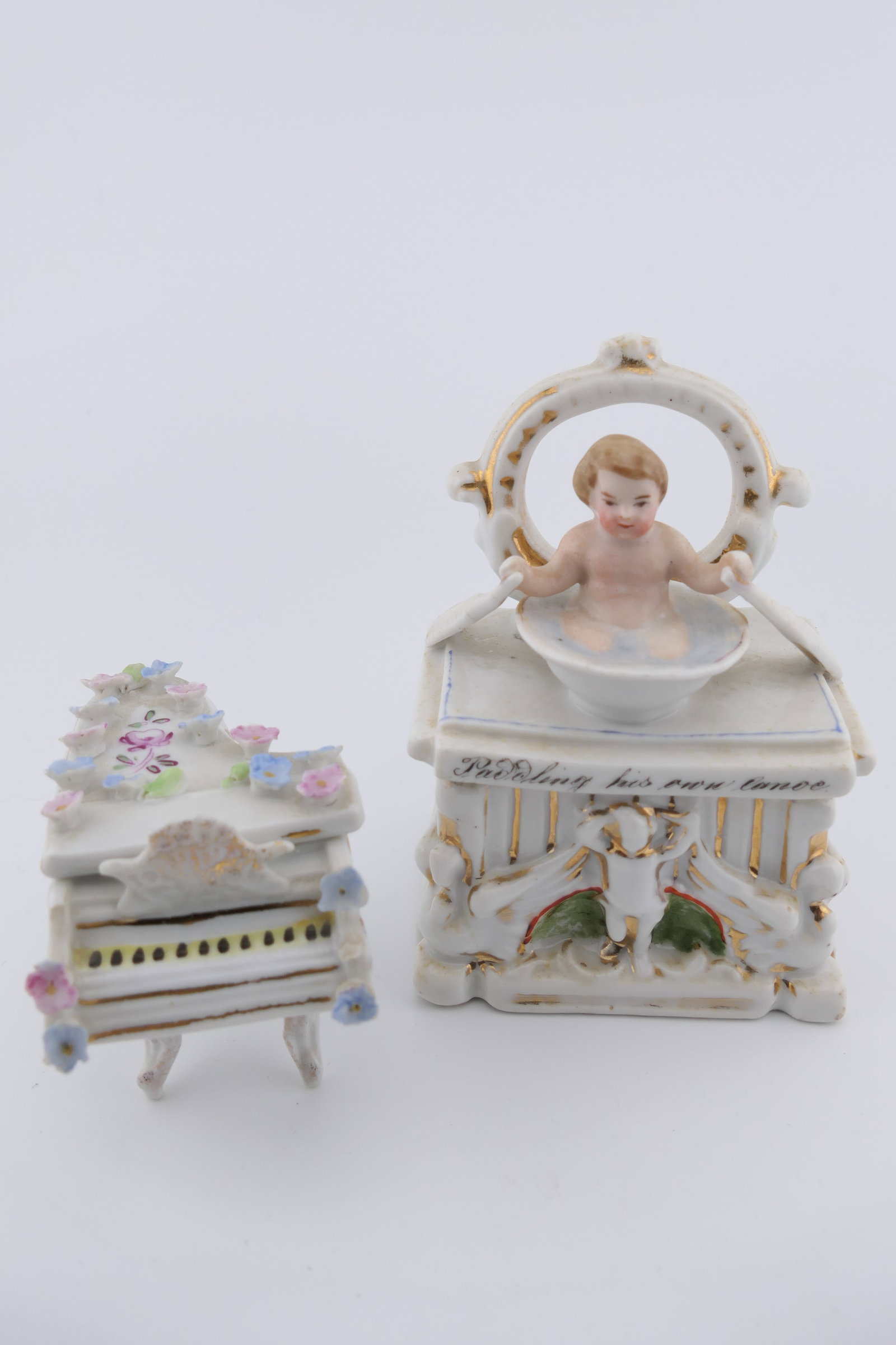 Two German Porcelain Trinket Boxes: Two German made porcelain trinket boxes including an antique circa 1900 Conta & Boehme box fashioned to look like a child in a wash basin, alongside a miniature piano with floral accents. The