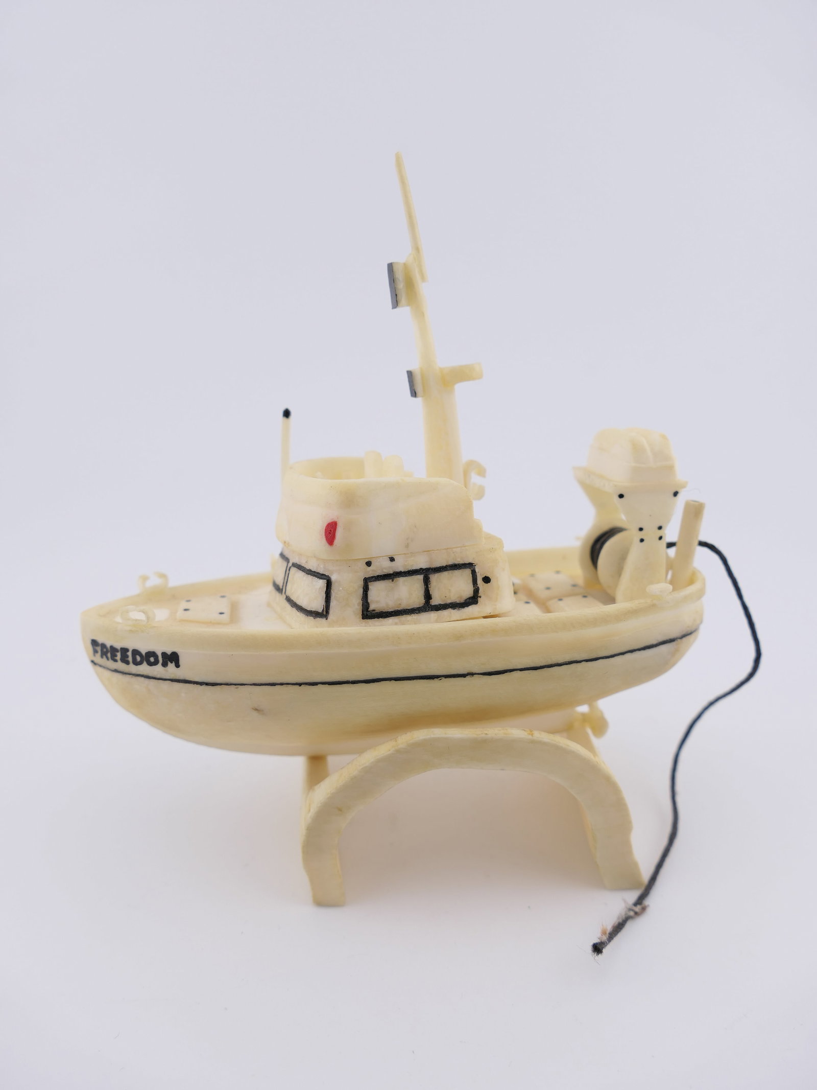 Hand Carved Bone Boat Statuette: Hand carved statuette in bone, featuring a design of a fishing boat, painted on the side reading 'Freedom'. Marked on the back with artist initials "KTN.AK." Rests upon a separate carved bone stand. A