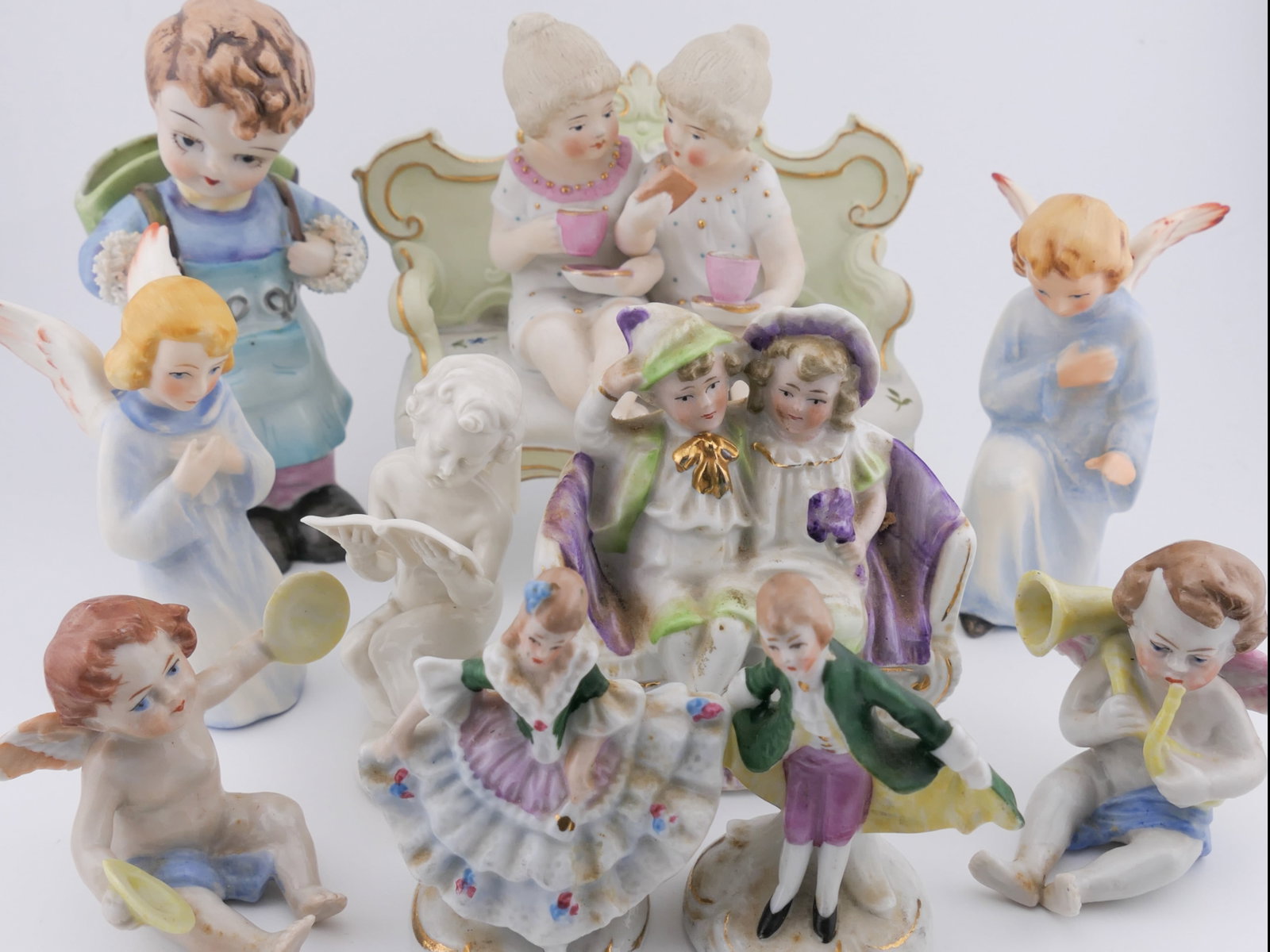 Assorted Porcelain Figurines: Assorted porcelain figurines including five marked Germany, two marked West Germany, and one marked Italy. Styles include a cherub reading, a bisque figurine of two girls sharing tea on a settee, two 