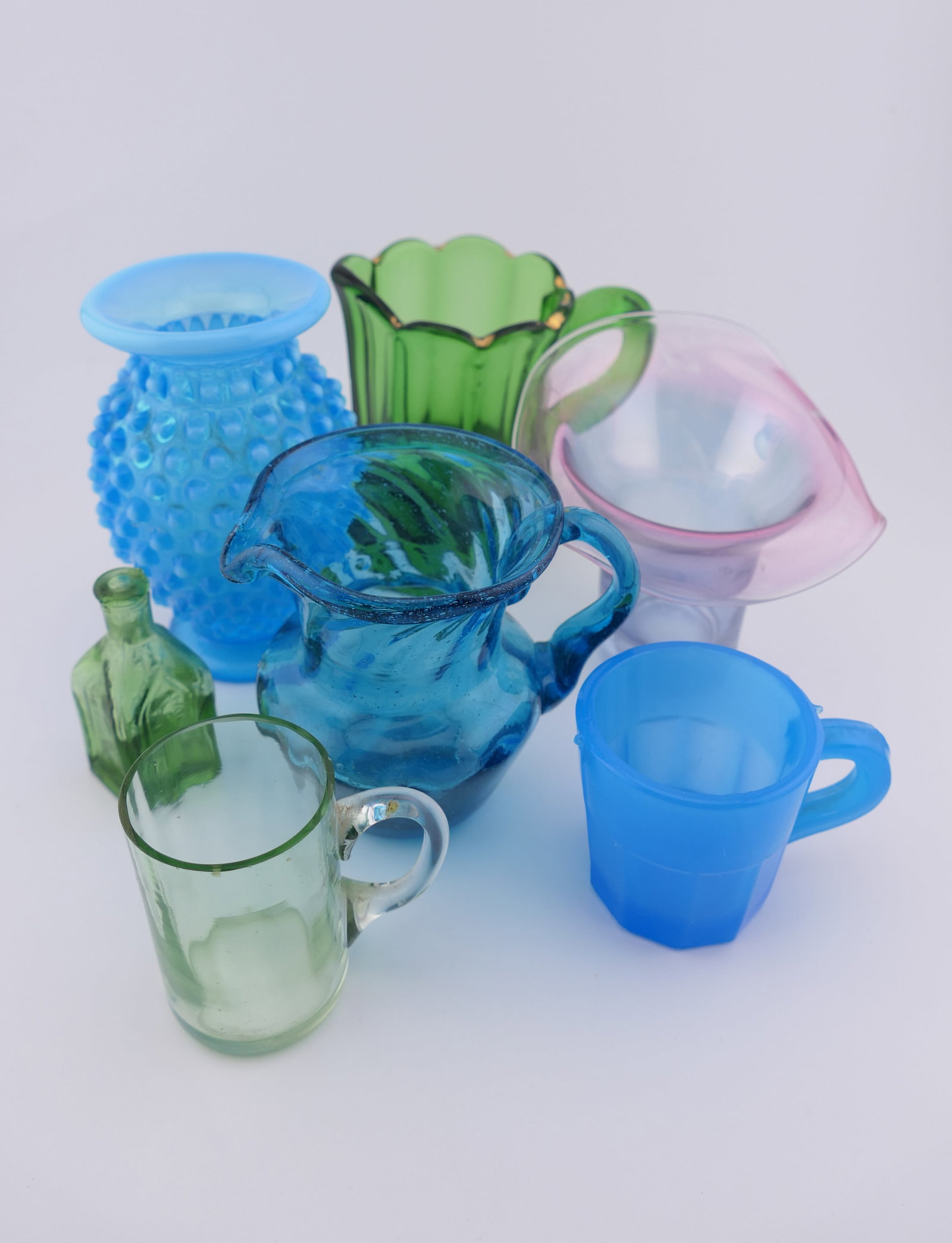 Vintage Accent Glassware: Vintage accent glassware including a blue hobnail vase, a blue swirl glass pitcher, a green glass pitcher, a pink and blue iridescent vase, a small vaseline glass mug, and a Cathedral Brand Chief