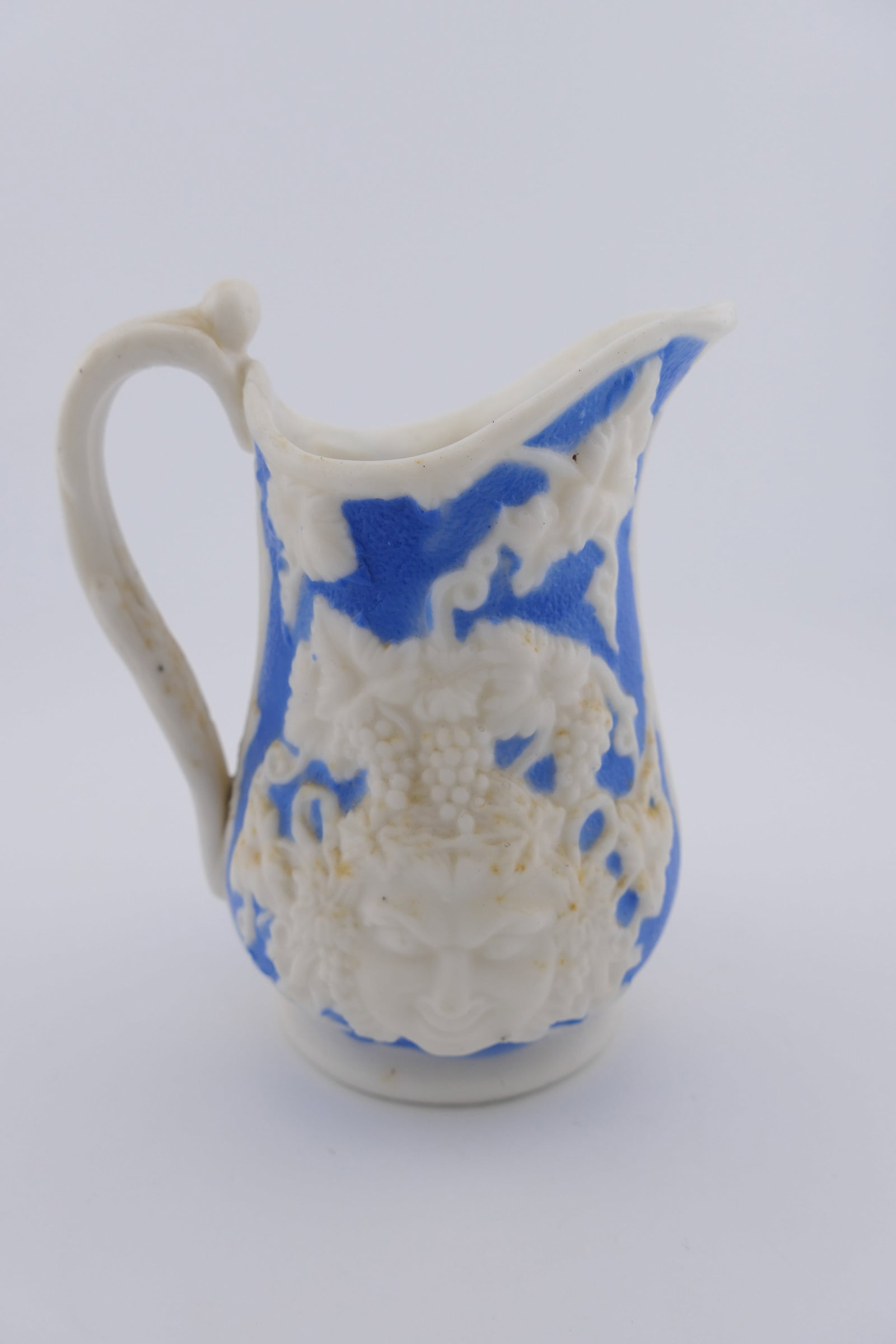 Small Neoclassical Relief Jug: Small neoclassical jug featuring a cream relief motif of grape clusters and vines on a blue field that frame a Bacchus or Dionysus style mask. The interior is glossy, the exterior is matte. Very good 
