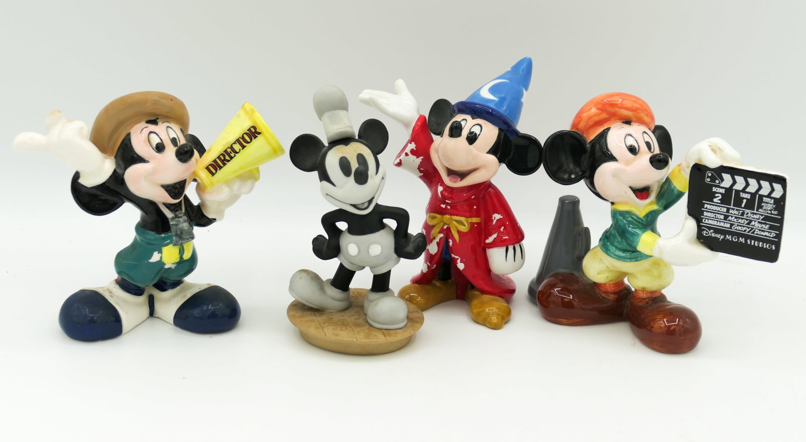 Mickey Mouse Ceramic Figurines: Four Mickey Mouse ceramic figurines including Director Mickey with megaphone, Director Mickey with film slate, Sorcerer Mickey and Steamboat Willie. Director Mickey figures show significant paint