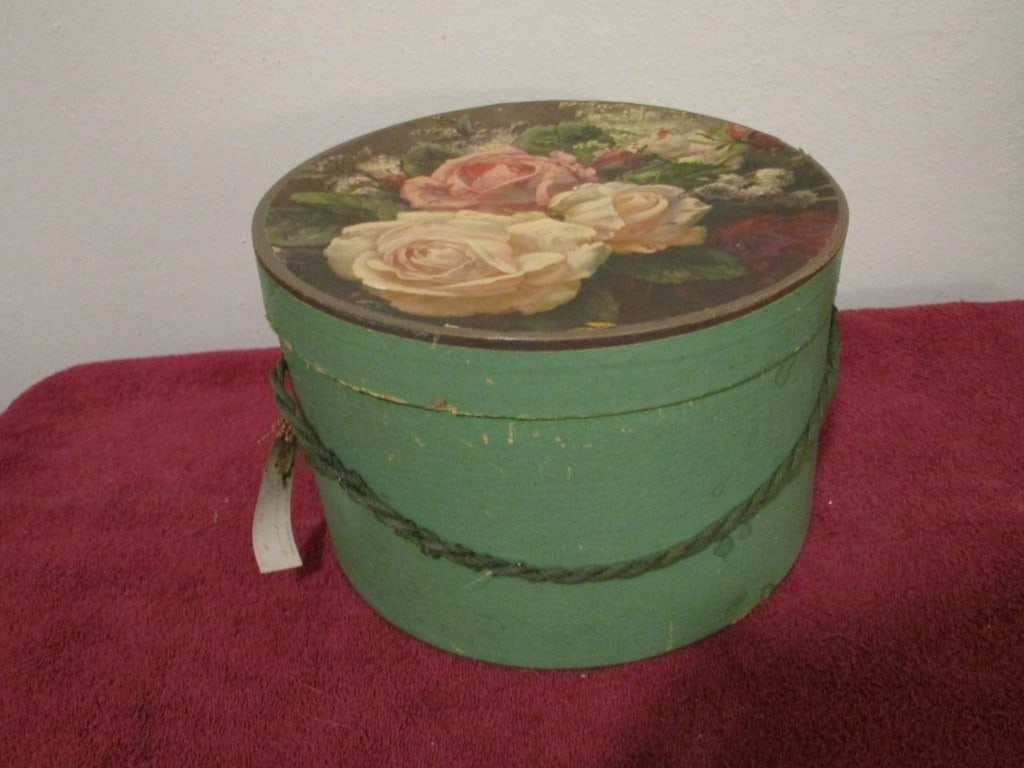 1930s Hat Box, Green Floral Design on Lid: 1930s Hat Box, Green Floral Design on Lid. Original rope and Christmas tag. Some loss on rim, some discoloration, water marks, and paint / paper loss. No cardboard separation. Meas 7"h x 11"r.