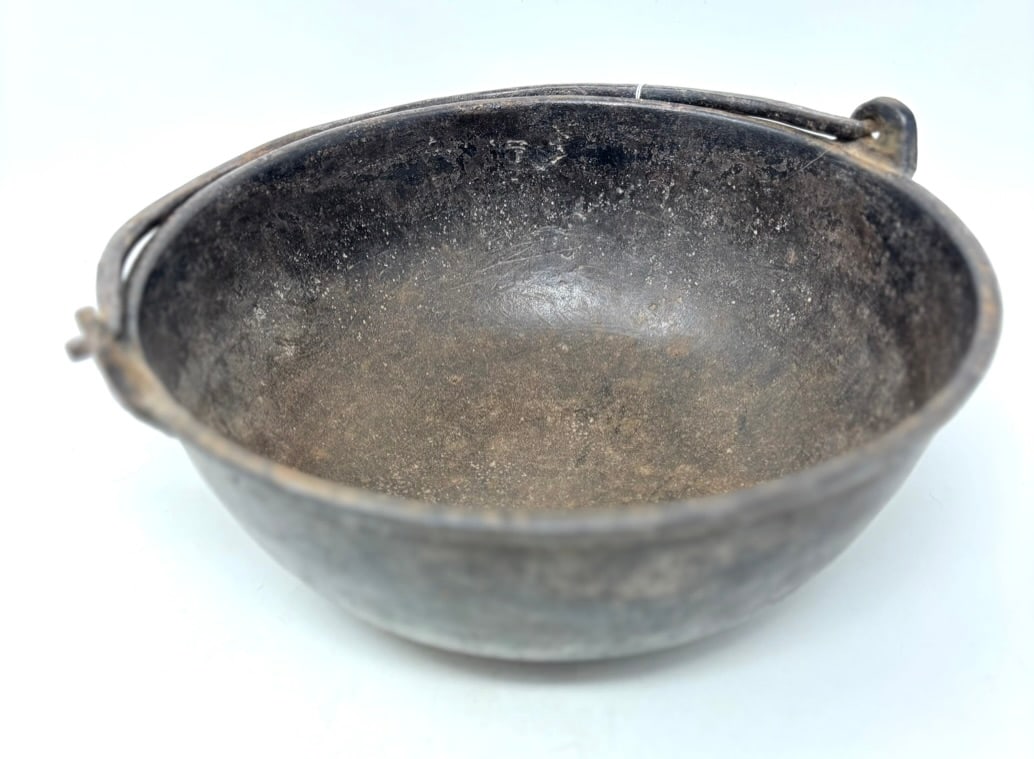 Early Cast Iron Pot (Cauldron): An early cast iron pot (cauldron), 9 1/2” dia. x 4” high having a flat bottom, rimmed top, line casting gate on bottom, flat cast ears, and a hand forged bail handle. Called a “Cowboy