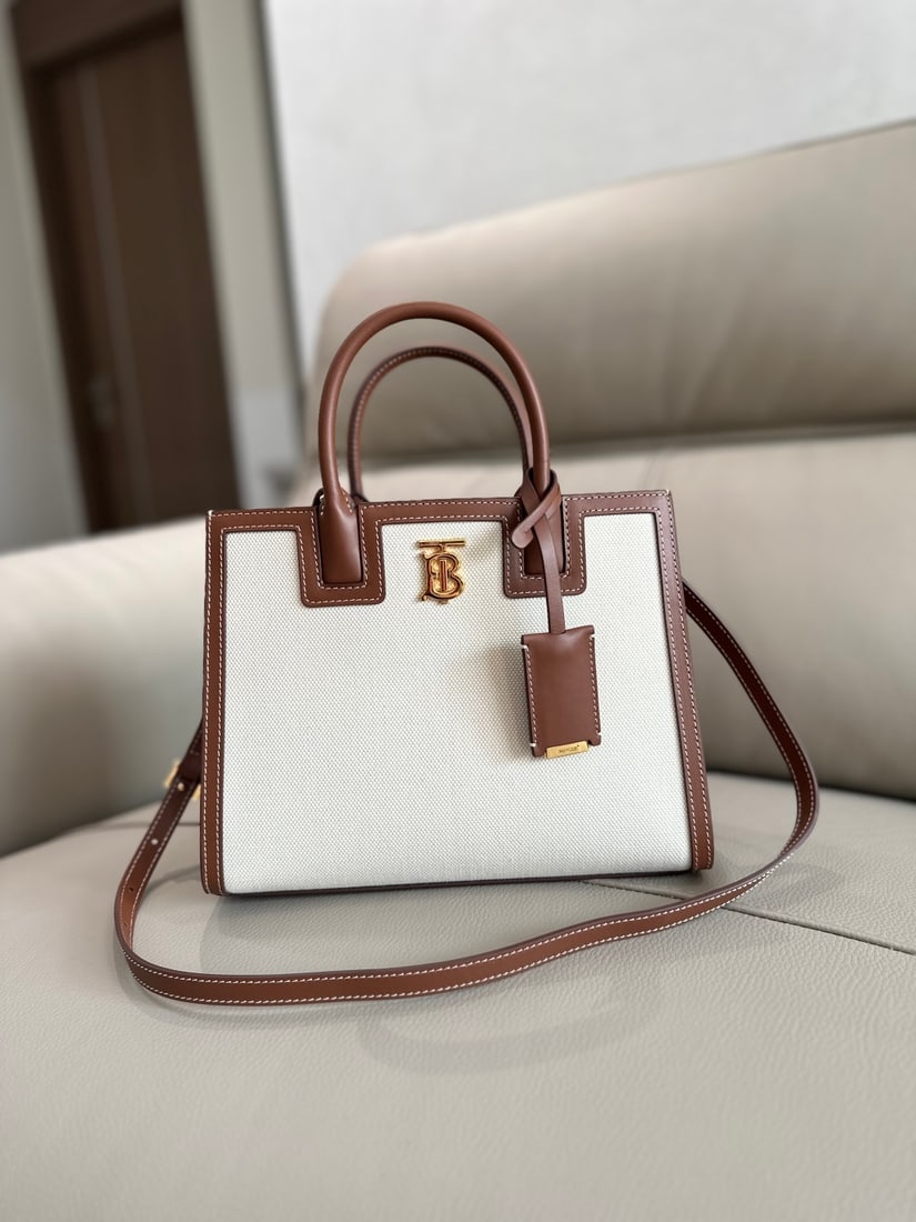Burberry: This Burberry tote bag is crafted from Italian-tanned leather and canvas, adorned with the Burberry logo. It features a calfskin lining with a mirrored charm. Dimensions: 27 x 11 x 20 cm. Decoration: 