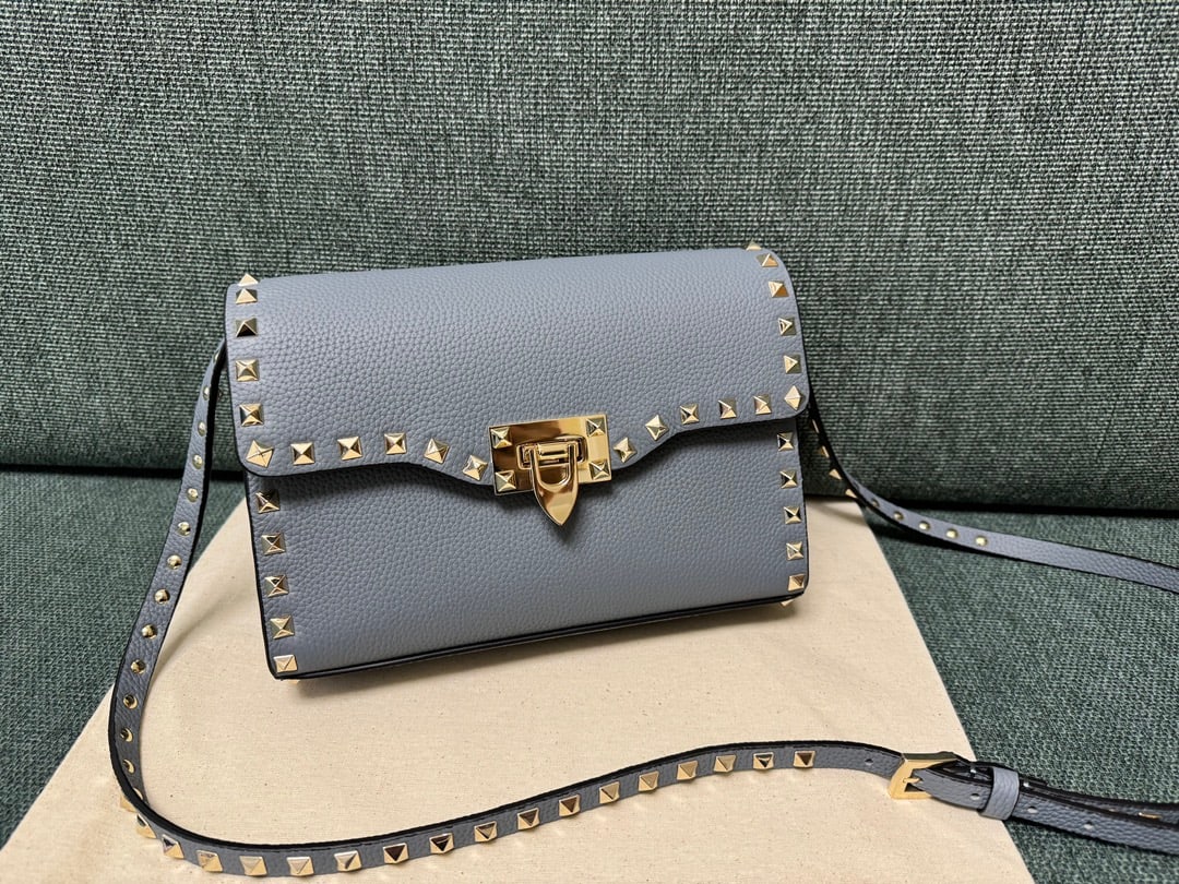 Valentino: This Valentino messenger bag in grey-blue with tumbled gold studs features a simple design, an adjustable shoulder strap, and one interior slip pocket. It's made of plain-grain cowhide with a natural 