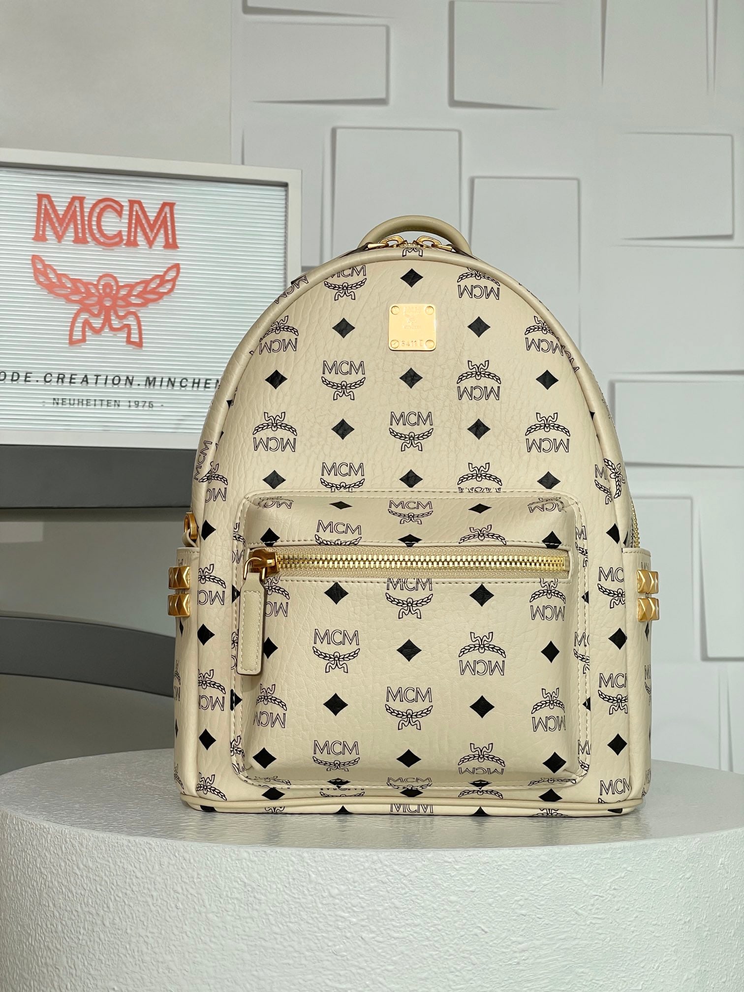 MCM Strak Visetos Off-White Small Studded Backpack: MCM Strak Visetos Beige Small Studded Backpack. This new colorway backpack features a well-structured design that redefines the vintage feel of hardware. The distressed, brushed gold finish exudes a p