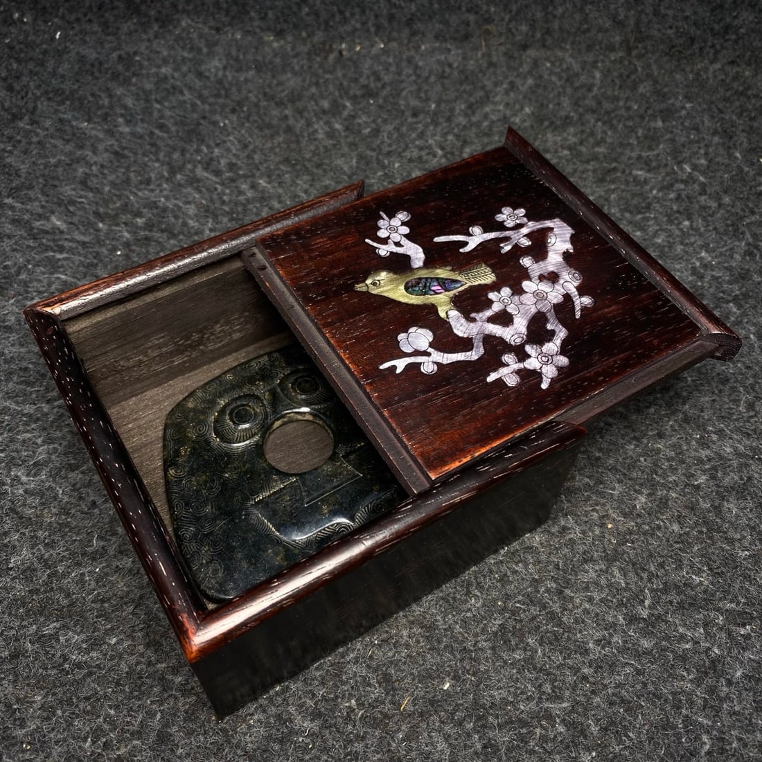 A precious Hetian jade disc is housed inside a rosewood box inlaid with mother-of-pearl.: A rosewood box inlaid with mother-of-pearl contains a precious Hetian jade bi disc. The bi disc measures 9.8 x 8.2 x 0.7 cm (length x width x thickness). The box measures 12 x 12 x 6 cm (length x widt