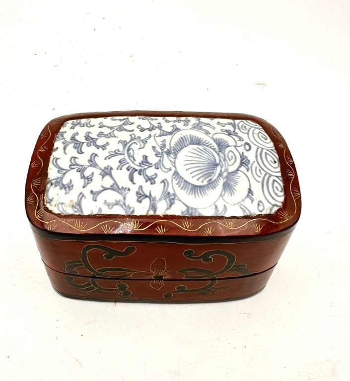 A rectangular two-tier lacquered wood box inset with a blue and white porcelain panel finely: A rectangular two-tier lacquered wood box inset with a blue and white porcelain panel finely painted with scrolling peony and lotus flowers. The body finished in a red-brown lacquer with gilt and blac