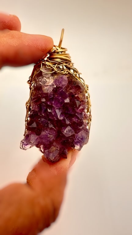 A striking natural amethyst crystal cluster pendant, featuring deep purple druzy crystals with: A striking natural amethyst crystal cluster pendant, featuring deep purple druzy crystals with good natural luster and faceted terminations. The stone is hand-wrapped in gold-tone wire, forming an org
