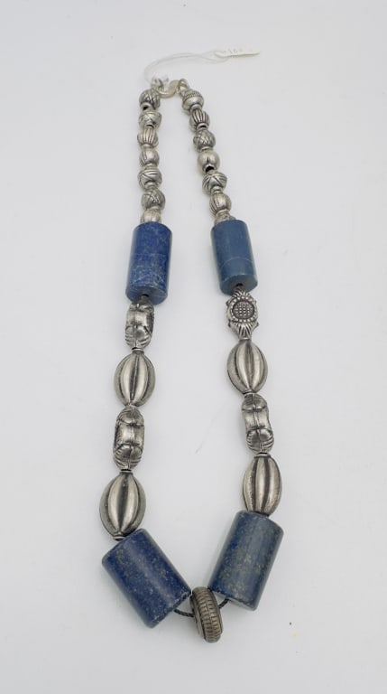 A bold and distinctive necklace composed of large cylindrical lapis lazuli beads in deep blue: A bold and distinctive necklace composed of large cylindrical lapis lazuli beads in deep blue hues, accented by ornate silver beads of various forms, including ribbed ovals, spherical, and stamped flo