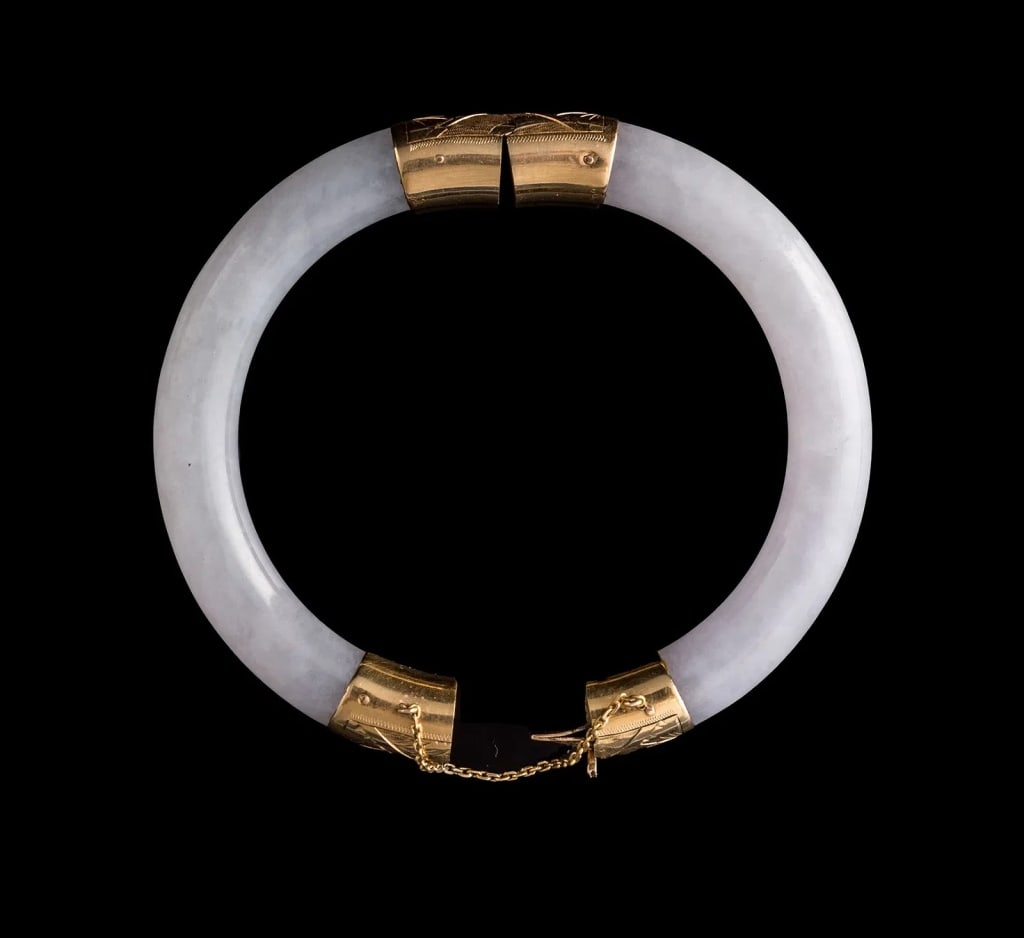 CHINESE LAVENDER JADEITE AND 14K GOLD HINGED BANGT A polished Chinese lavender jadeite bangle: A polished Chinese lavender jadeite bangle bracelet, constructed in two sections and fitted with 14K gold mounts, engraved decoration, hinged fittings, clasp and safety chains.Material: Jadeite, 14K g