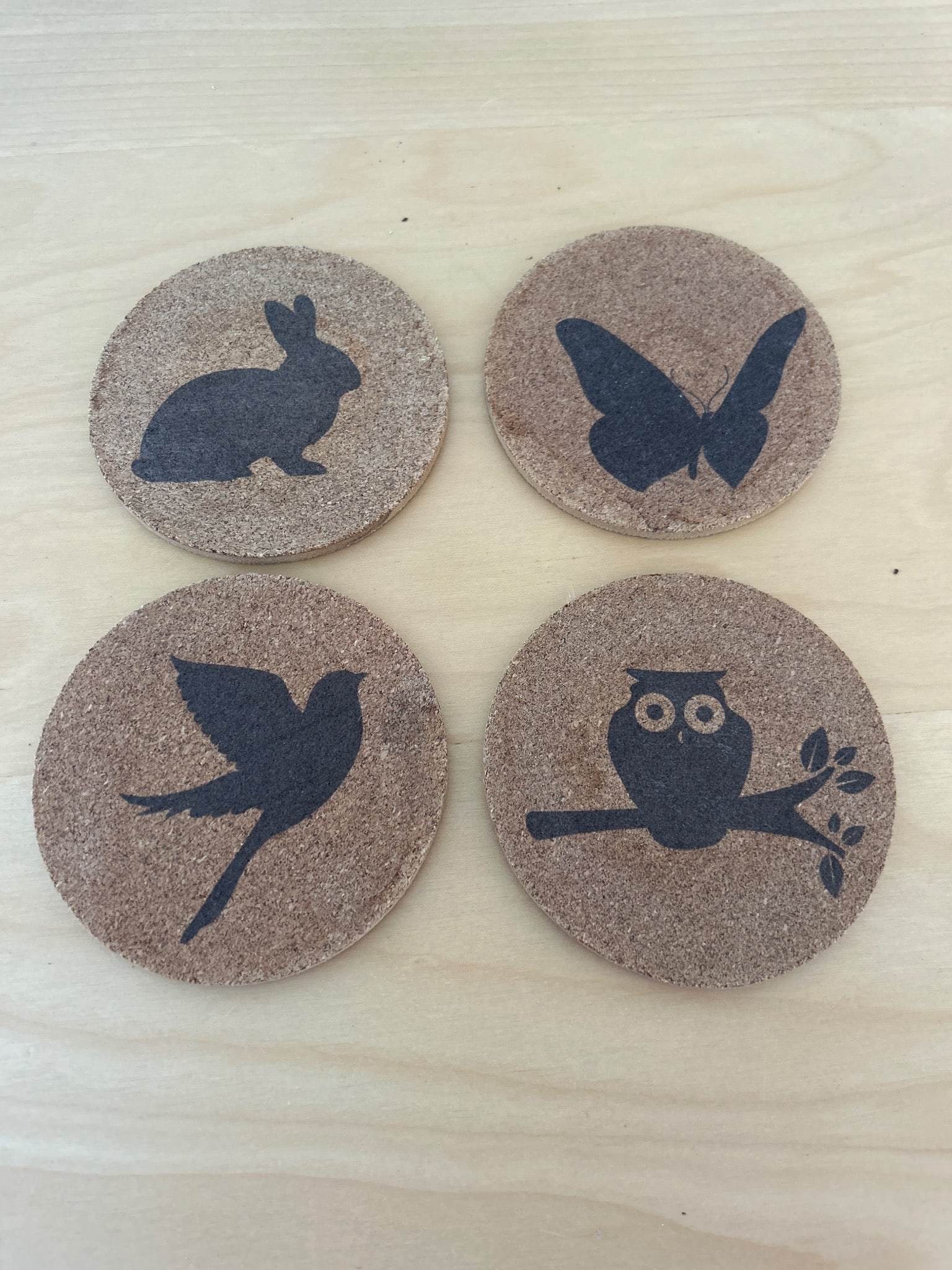 4 Mainstays Cork Coasters With Animal Silhouettes: Four cork coasters by Mainstays featuring black silhouettes of a rabbit, butterfly, bird, and owl on smooth cork surfaces. Designed for table surface protection and decorative use. Each coaster has a